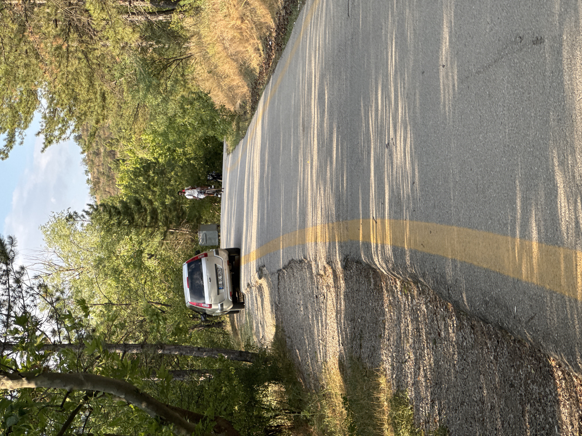 The image depicts a paved road winding through a forested area, likely on the outskirts of Gölbaşı, Turkey. A silver SUV is parked on the gravel shoulder to the left of the road. Further up the road, a person is riding a bicycle. The road is marked with a yellow line. The surrounding environment is characterized by lush green trees, with some dry grasses and bushes also visible, especially on the right side of the road. The lighting suggests it is late afternoon or early evening, casting long shadows across the road.