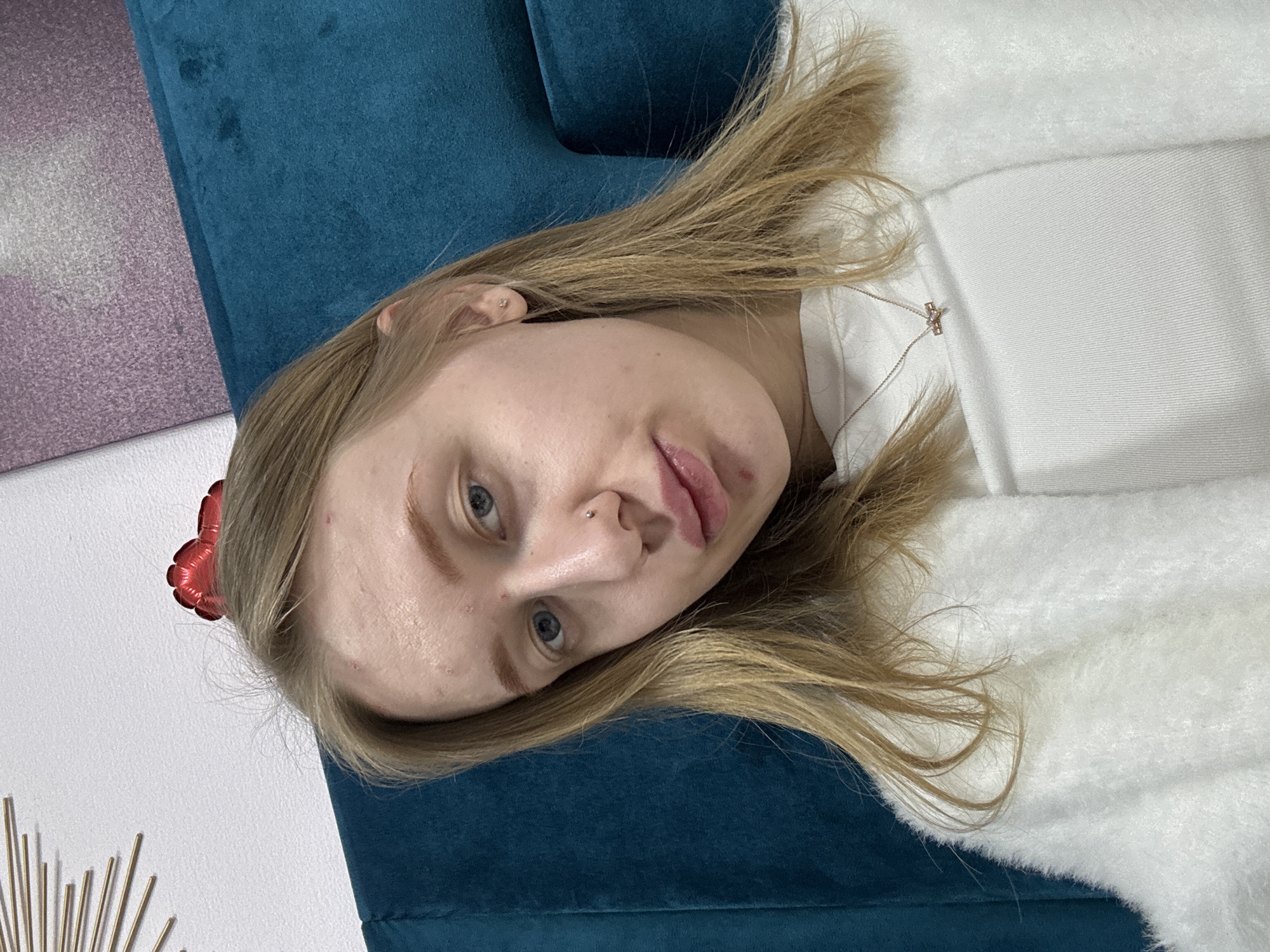A blonde woman with fair skin and blue eyes lies on a blue velvet couch. She wears a white, fluffy sweater and a silver necklace with a cross pendant. A small, red heart-shaped balloon is visible near her head. The setting includes a glimpse of a purple wall and a gold, sunburst-shaped decoration. The location information is not discernible from this image.