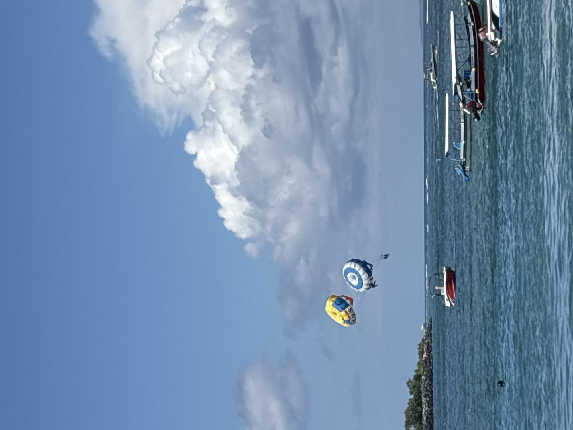 The image depicts a coastal scene in Tanjung Benoa, Indonesia. The sky is predominantly blue with large, puffy white clouds. Two parasailers are suspended in the air; one has a yellow and red parachute, while the other has a blue and white parachute. The ocean is visible on the right side of the image, with several boats and other watercraft. A strip of land with some vegetation is present between the ocean and the skyline.