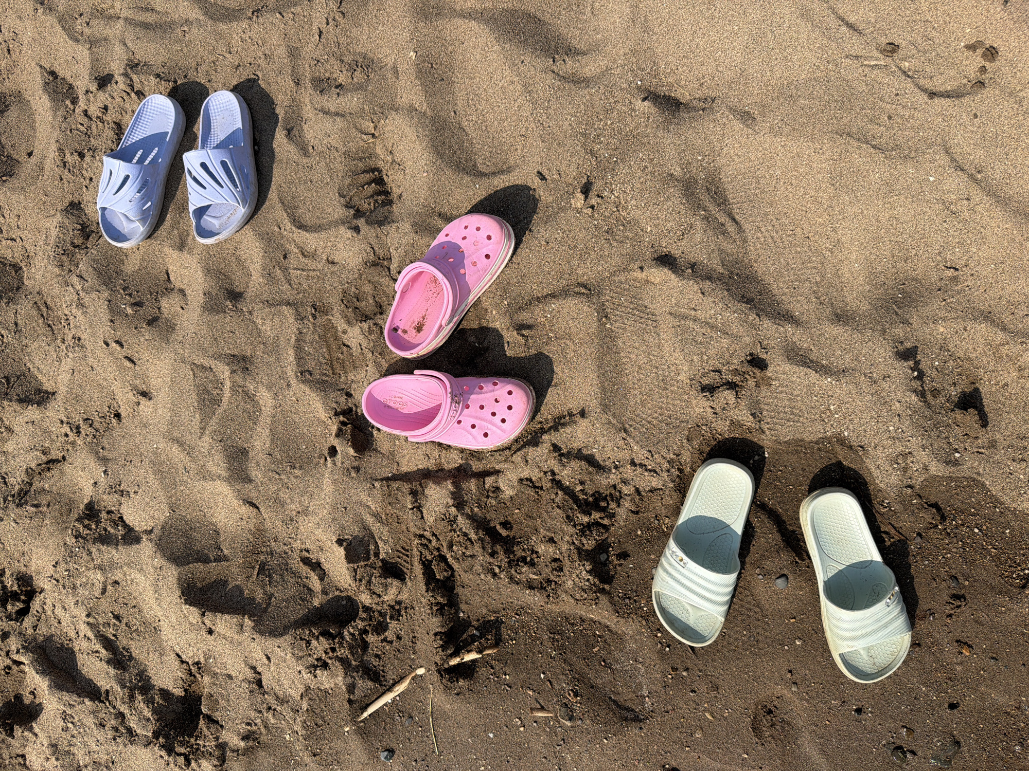 An overhead shot shows several pairs of sandals and crocs scattered on dark, damp sand. The footwear includes a pair of light blue slides, a pair of pink crocs, and two pairs of white slides. The sand has numerous footprints and some scattered twigs and pebbles. The setting is a beach in Krasnyi Bor, Russia.