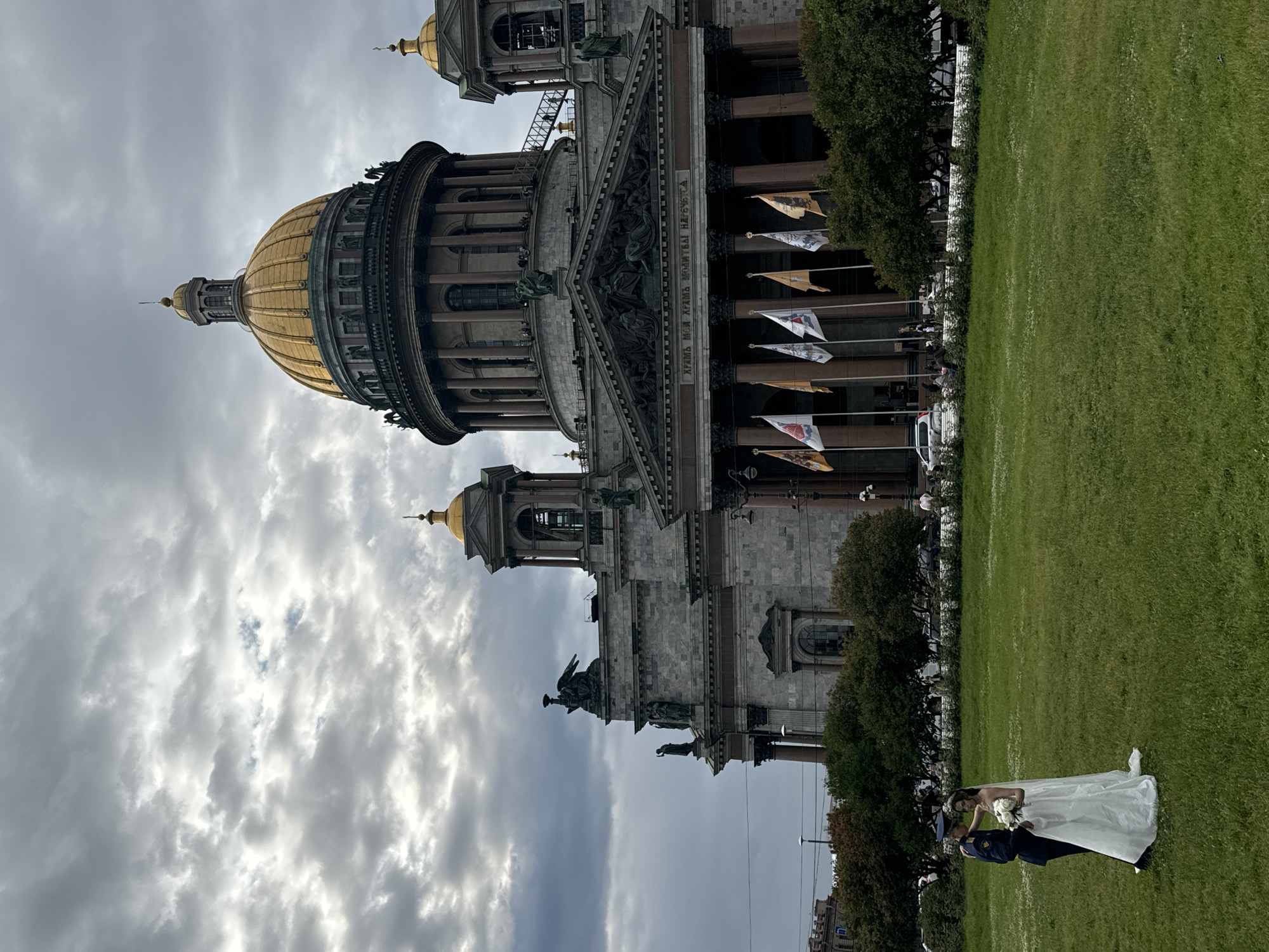 The image captures Saint Isaac's Cathedral in Saint Petersburg, Russia, under a cloudy sky. In the foreground, a bride in a white wedding dress and a groom in a dark uniform stand on a well-maintained green lawn. The groom is placing his hand on the bride's shoulder, and she is holding a bouquet of white flowers. Behind them, the grand facade of Saint Isaac's Cathedral is visible, featuring its iconic golden dome, columns, and ornate architectural details. Several flags are displayed along the front of the cathedral. The area around the cathedral appears to be a public space, with trees and a street with vehicles in the background.