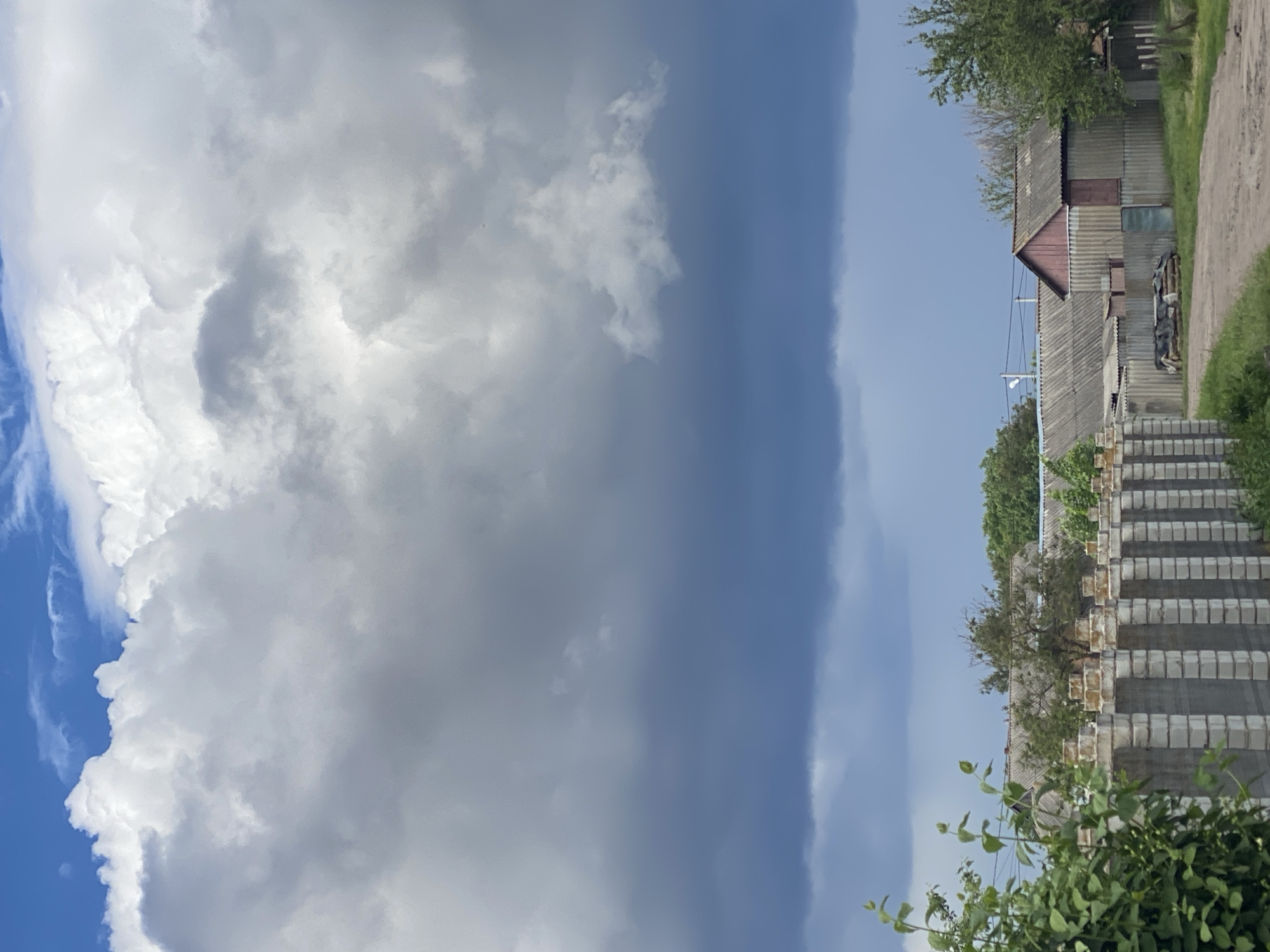 The image depicts an outdoor scene in Ustyvytsia, Ukraine. Dominating the left half is a large expanse of cumulus clouds against a blue sky. To the right, the scene transitions into an area with buildings and vegetation. A gray corrugated metal building is visible, with a brown gable roof section. A set of concrete stairs leads upwards, flanked by trees and greenery. In the foreground, foliage partially obscures the view. A dirt road runs alongside the building.