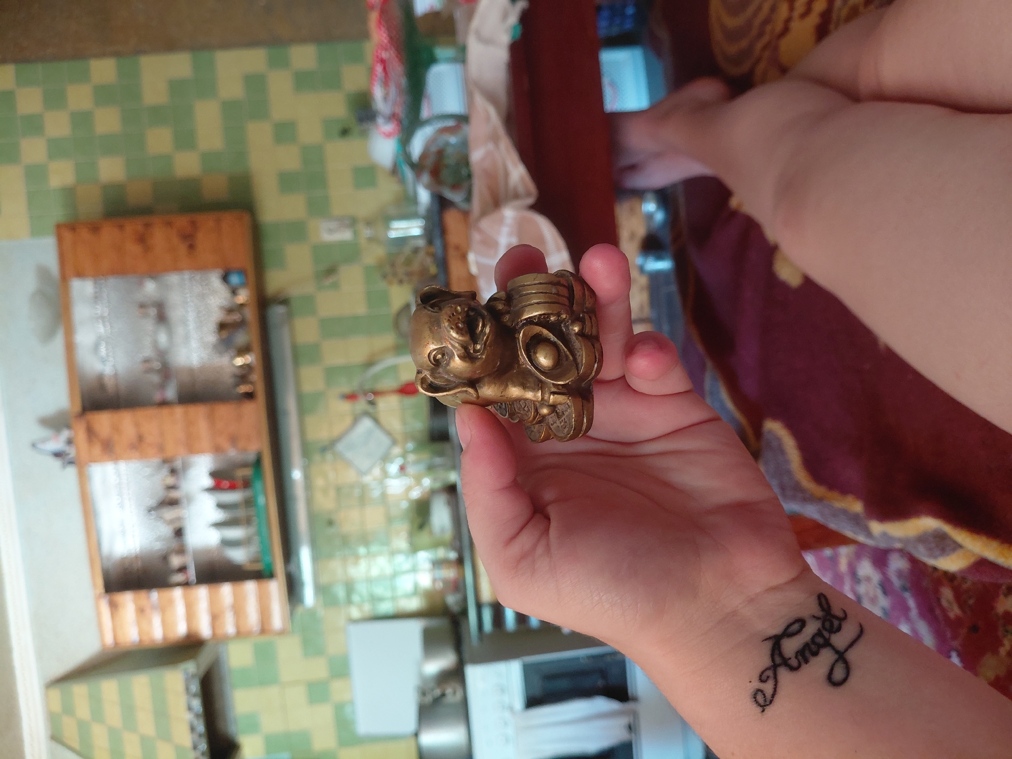 The image shows a close-up of a person's hand holding a small, ornate brass figurine. The figurine appears to be a stylized animal, possibly a cat or a dog, with large eyes and a textured body. The person's wrist is also visible, with a tattoo that reads "Angel" in script. The background is a tiled floor with a pattern of green and yellow squares, and some wooden shelves filled with small items. There is also a glimpse of a person's legs in the upper right of the frame, suggesting a seated or reclining position. The lighting suggests it is daytime. The setting appears to be indoors, likely a domestic environment in Marynivka, Ukraine. The image is taken from a slightly overhead angle.