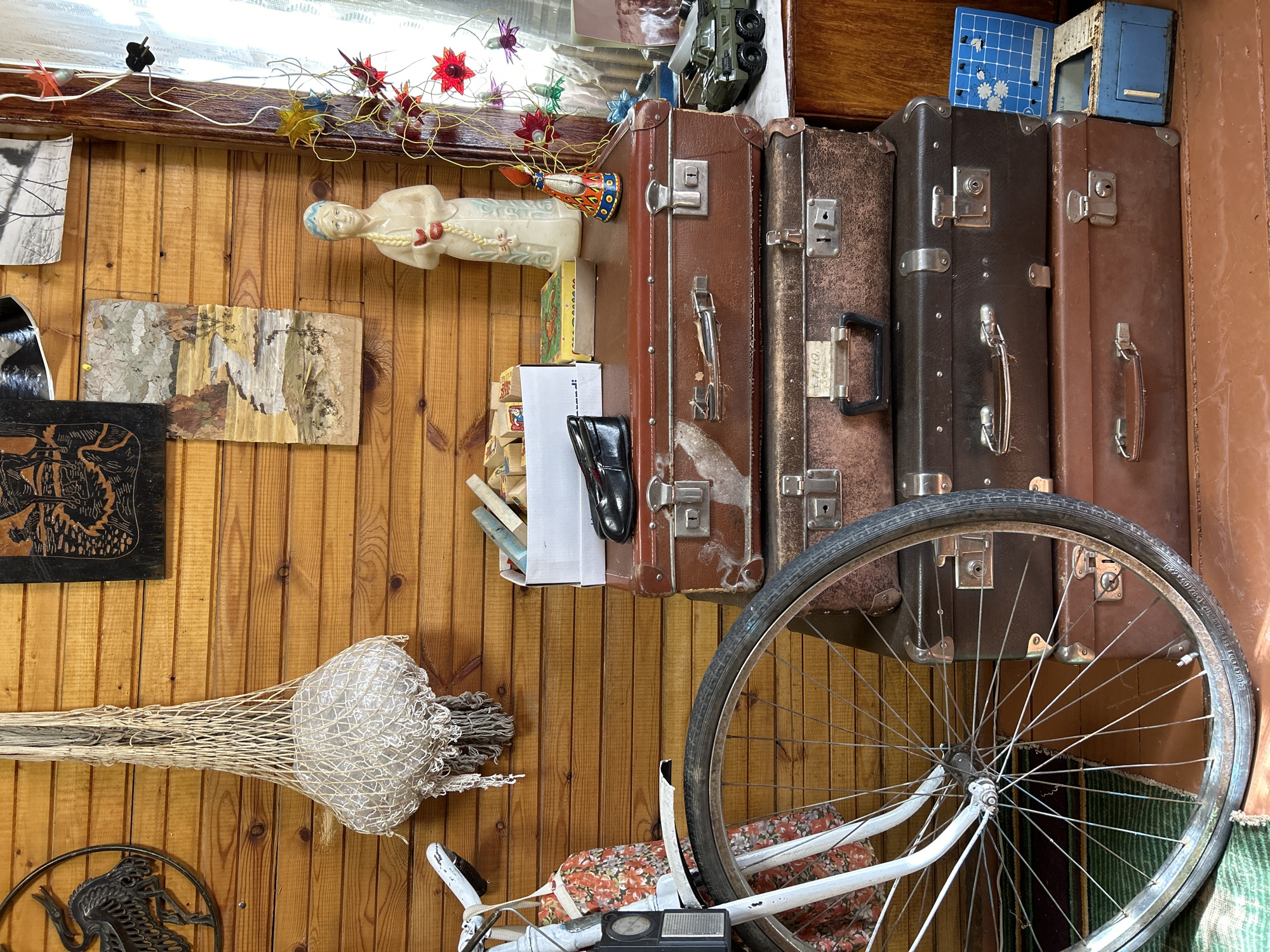 The image presents an eclectic, cluttered interior space with a strong emphasis on vintage and repurposed items. The walls are lined with vertical wooden planks. An assortment of decorative objects and personal effects are hung on the walls including a sculpture of a swimming person, a piece of driftwood, black-and-white photography, and a woven straw object. A row of vintage suitcases is stacked on a shelf, some displaying metal clasps. A partially visible bicycle, with its front wheel and handlebars in focus, occupies the lower right of the frame, its seat covered in a floral-patterned fabric. The specific location within the city of Moscow, Russia, cannot be identified with certainty from the visual information available.