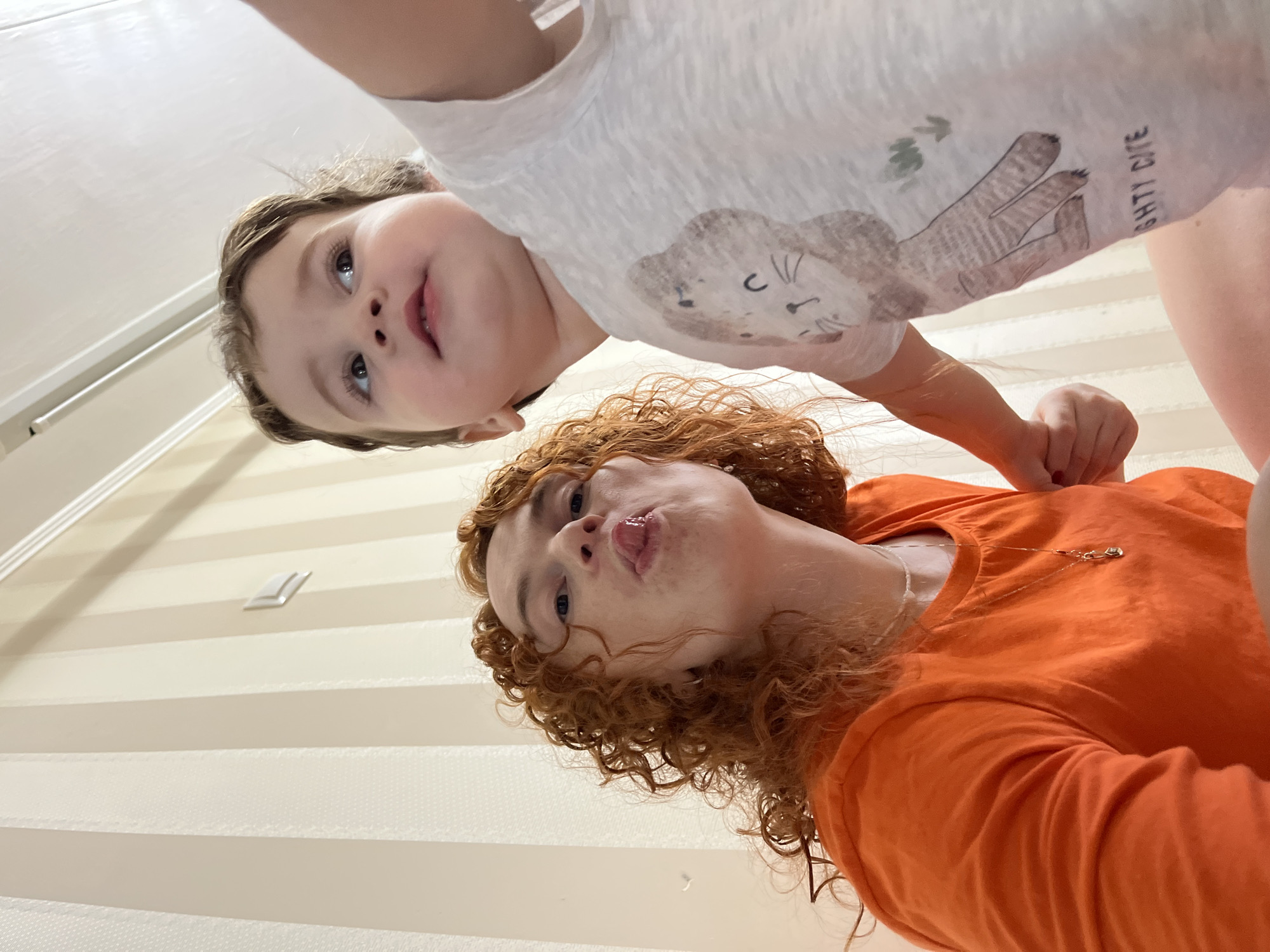 A low-angle shot shows a woman and child. The child, wearing a white t-shirt with a cat design, is in the foreground. The woman, with curly red hair and wearing an orange t-shirt, is behind the child, sticking her tongue out. The background consists of a wall and a light fixture. The location is not specified, but the prompt asks about the city of Chaikovsky, Russia.
