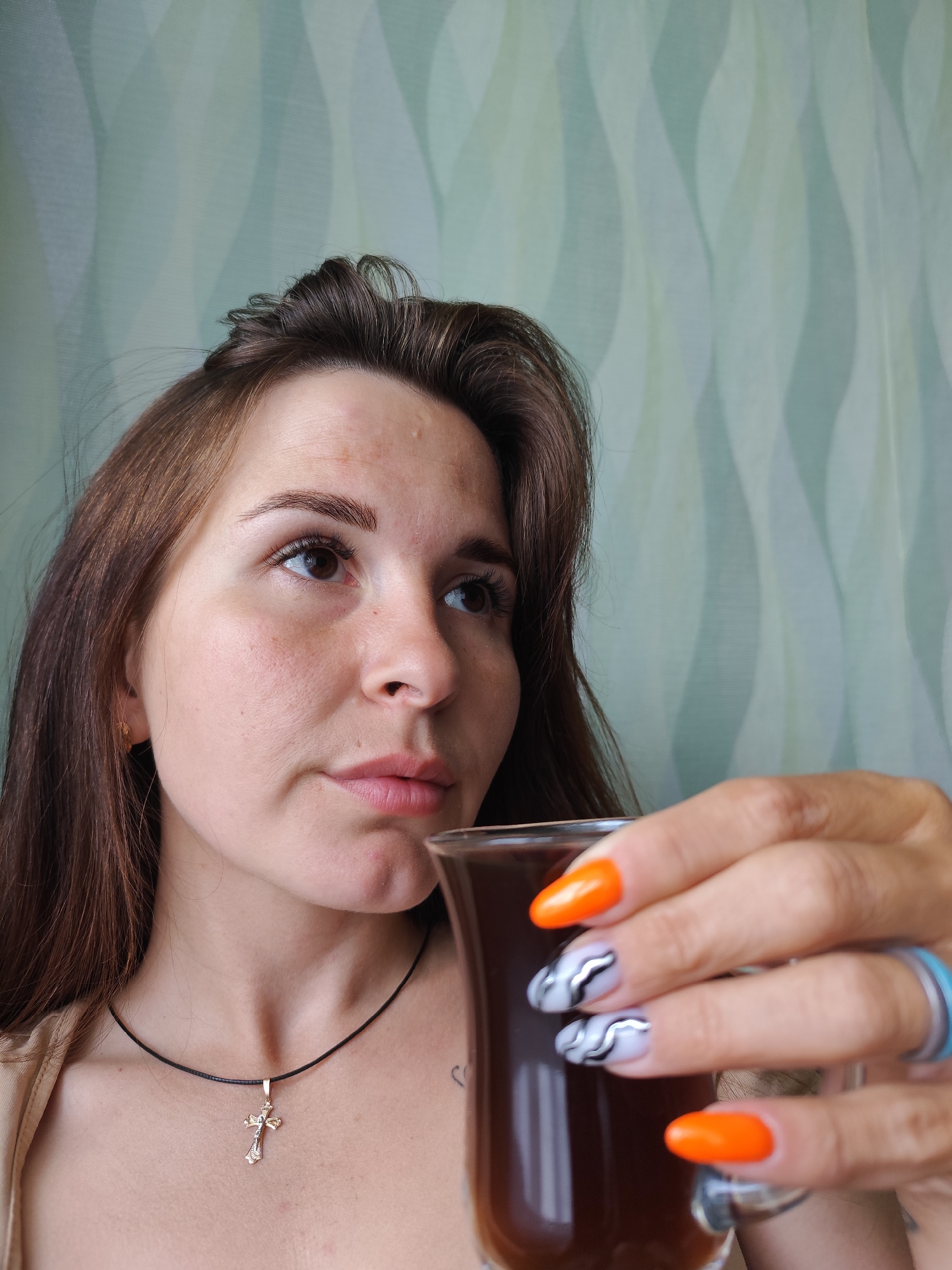 The image portrays a young woman, likely in her 20s or early 30s, captured from the chest up. She is positioned slightly to the left, looking towards the upper right of the frame with a thoughtful or pensive expression. Her long, dark brown hair frames her face, and her eyebrows are dark and well-defined. She is wearing a light, possibly beige, top, and a black cord necklace with a golden cross pendant rests on her chest.
Her right hand is prominent in the foreground, holding a clear, tall glass containing a dark beverage, possibly coffee or tea. Her nails are distinctively decorated: some are painted a bright, solid orange, while others feature a lighter base with intricate black and silver wavy designs. A light blue ring is visible on her right ring finger.
The background consists of a soft green and light blue patterned surface, which appears to be either wallpaper or a curtain with subtle, undulating vertical stripes. The lighting suggests natural daylight, illuminating the scene. The overall ambiance is calm and personal. There is no visible text in the image. While the location context is given as Foki, Russia, no specific visual cues within the photo directly indicate this geographical detail.