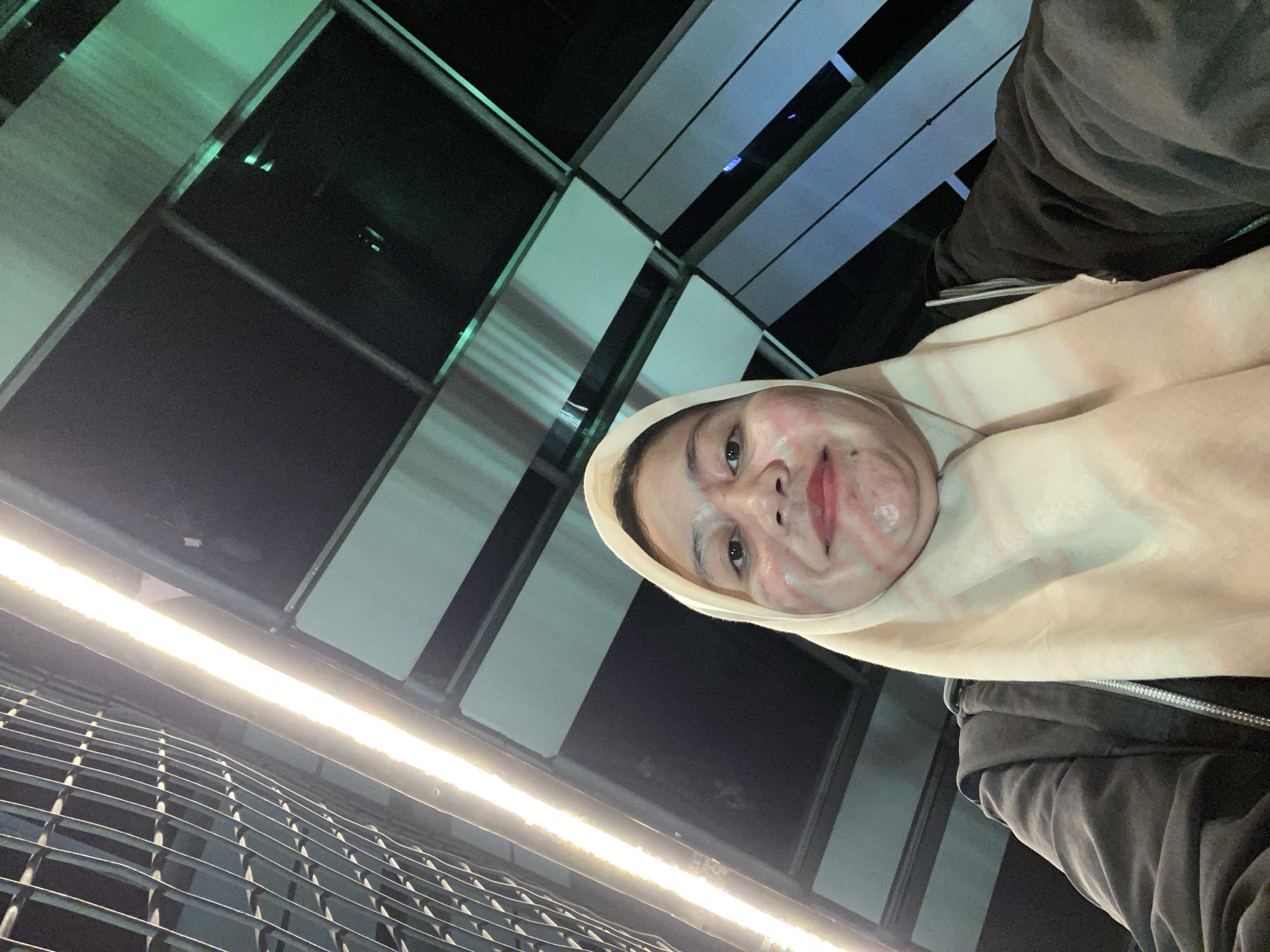 This media file shows a selfie of a person in Banjarbaru, Indonesia. The person is wearing a light-colored hijab and a dark jacket. Their face is partially visible, with a playful expression. The background features an abstract architectural design with a combination of dark and light-colored panels, possibly part of a building or an outdoor structure. A long, bright light fixture is visible in the lower part of the frame, illuminating a metal grate or railing in front of it. The lighting and overall composition suggest it might be evening or nighttime. There is no visible text in the image.