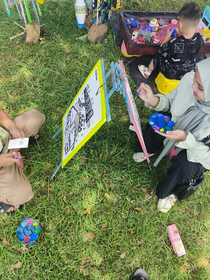 The scene takes place in a grassy park in Majalengka, Indonesia, located at latitude -6.846 and longitude 108.204. A woman, wearing a green headscarf and a black skirt, is sitting on a stool, painting on an easel. The easel is yellow and features a black and white drawing. She holds a paintbrush in her right hand and a blue and purple circular toy in her left. A young boy, wearing a black and white shirt with a graphic design, sits on a yellow stool, facing away from the camera. He is playing with a toy truck. A man wearing a beige shirt and brown pants sits in the foreground, holding a small piece of paper in his hand. A red and white container, containing various items, sits on the ground to the right of the woman. A blue and purple toy, a yellow plastic container, and several items of unidentified objects lie on the ground in front of the woman and the boy. The environment consists of lush green grass and scattered leaves.
