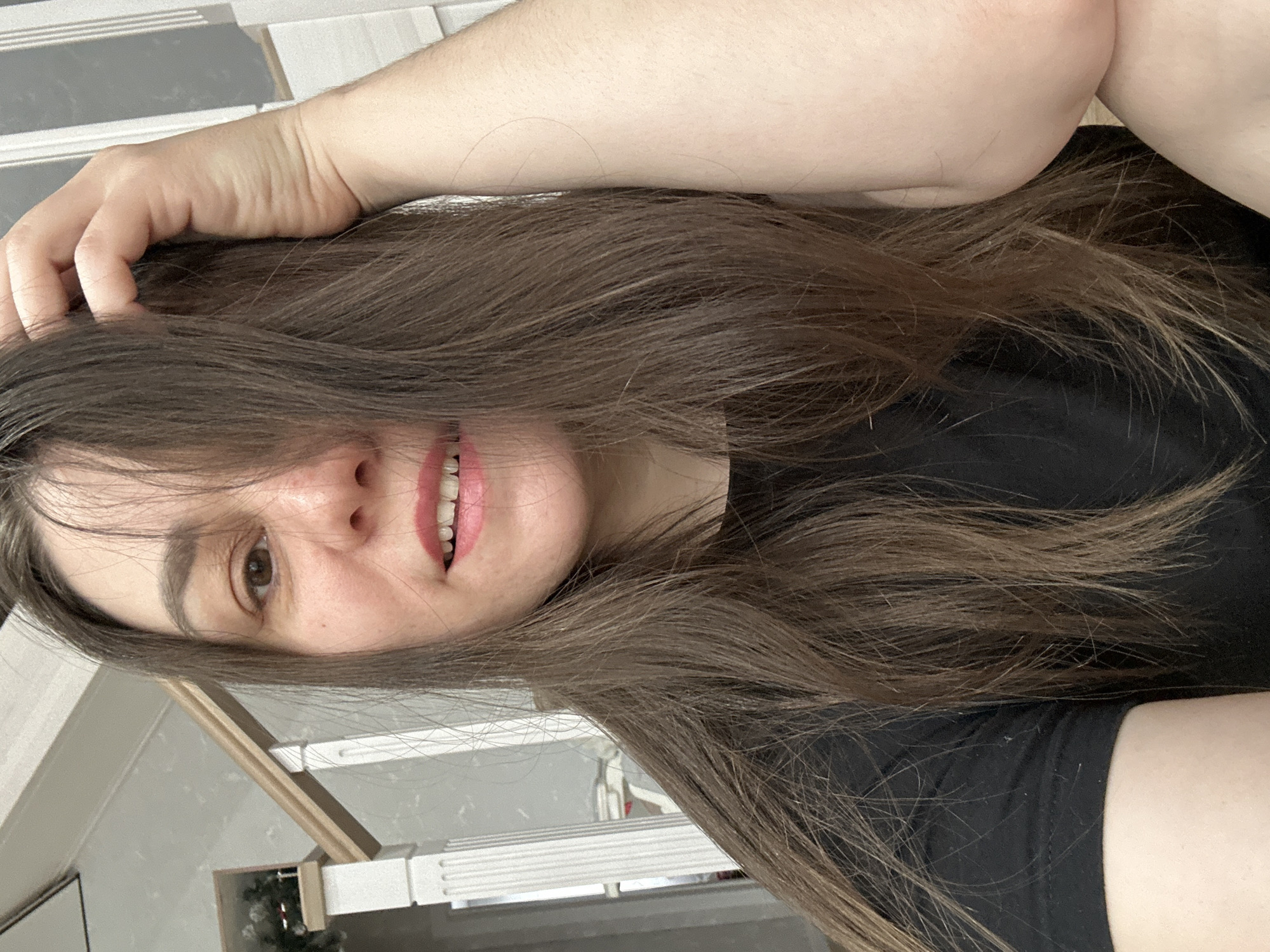 The image is a close-up selfie of a woman with long brown hair. She is smiling and her hand is touching her hair. She is wearing a black top. The background is blurred, but appears to be an indoor setting. It is not possible to determine the location in the city of Chaikovsky, Russia from this image.