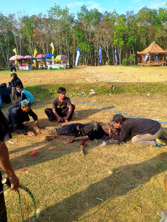 This image depicts an outdoor scene in an open field, potentially in the city of Kuto, Indonesia, under clear daylight.
In the mid-ground, a male subject lies supine on the dry, partially grassy ground, his clothing and exposed skin, including his face, covered in dirt. He wears a dark t-shirt with light-colored text and dark pants. Near his left hand lies a red-handled object with a chain, resembling a whip. Another male subject kneels beside the supine individual's head, leaning closely, appearing to attend to him or administer aid. This individual wears a dark long-sleeved shirt, light pants, a dark baseball cap, and sneakers. A third male subject kneels near the supine individual's feet, holding one of his lower legs. He wears a dark long-sleeved shirt and a patterned head covering. A fourth male subject sits cross-legged directly behind the supine individual, observing the scene. He wears a dark t-shirt and dark pants. A thin blue rope or cord extends across the ground to the right from the area near the seated individual. Various small pieces of debris, including plastic cups, are scattered on the ground.
In the background to the left, several colorful vendor stalls with umbrellas (pink, yellow, blue, and multi-colored) are visible, with people present near them. To the right, a traditional wooden gazebo with a tiered, tiled roof stands. A dense line of trees forms the backdrop behind the stalls and the gazebo. Several blue and yellow flags or banners are positioned vertically in the field between the vendor area and the treeline. In the foreground, partially cropped on the left, a person's arm and hand are visible holding a braided, multi-colored rope or whip. The sun is positioned to the left, casting shadows to the right side of the scene.
