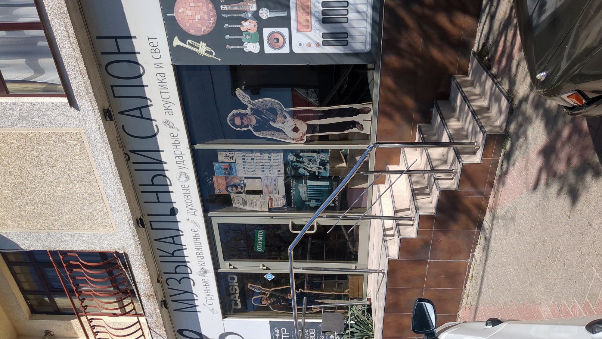 The image displays the exterior of a "Музыкальный Салон" (Musical Salon/Store) located in Gelendzhik, Russia. The building features a textured, light-colored facade with an entrance accessible via a set of tiled stairs with a metal handrail.
Above the glass entrance doors, a large sign reads "МУЗЫКАЛЬНЫЙ САЛОН" with smaller text below listing product categories: "струнные клавишные духовые ударные акустика и свет" (string, keyboard, wind, percussion, acoustics and light). The store's window displays feature large graphical representations of musical instruments, including guitars, a keyboard, drums, a microphone, a trumpet, and a disco ball. A life-sized cutout of a person playing an electric guitar is prominently displayed in the right window, and another guitarist cutout is partially visible in the left.
The left glass door has a "CASIO" logo, while the right door displays a green sign with "ОТКРЫТО" (OPEN). Various posters and advertisements are taped to the inside of the glass doors. The ground immediately in front of the store is covered in brown tiles, transitioning to a light-colored paved sidewalk on the right. Strong shadows from nearby foliage are cast across the stairs and sidewalk, indicating bright daylight, likely mid-morning or afternoon. A white car is partially visible on the far right, parked alongside the curb.