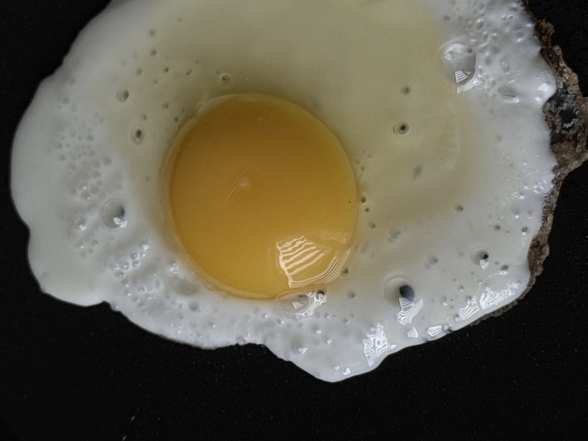 A close-up, top-down shot captures a fried egg on a dark, non-stick pan. The egg white is fully cooked and frothy, with small air bubbles dotting its surface. The yolk is bright yellow and appears to be partially cooked, with a glossy sheen. Reflections of a striped object are visible on the surface of the yolk and within the cooked egg white. The pan's surface is dark and textured, with some brown, burnt residue visible along the edge of the egg. The scene is entirely food-focused, with no people or environmental elements beyond the cooking surface. The image does not contain any contextual information about its location in Chaikovsky, Russia, or any other specific geographic area.