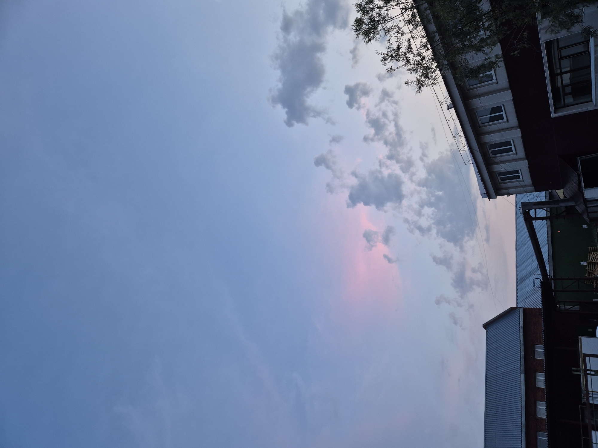 This image depicts the sky and buildings from a low-angle perspective, likely taken from a courtyard or between buildings. The scene is oriented such that the sky occupies the majority of the frame, with buildings visible along the right edge.
The sky is a soft gradient of blue, transitioning to lighter shades towards the horizon. Scattered clouds are present, some appearing as wispy streaks and others as more defined masses. There's a subtle hint of pink or purple in the clouds, suggesting either a sunrise or sunset. The presence of tree branches overhanging from the top right corner indicates a natural element in the urban environment.
The buildings appear to be multi-story structures, likely residential or commercial. The architectural style suggests a somewhat functional design, with visible windows and varied materials such as brick, metal siding, and what looks like concrete or rendered facades. The building in the foreground has a prominent ribbed metal siding.
The time of day is either early morning or late evening, as indicated by the soft lighting and the colors in the sky. The weather appears calm, with the clouds not suggesting any severe conditions. There are no people visible in the image. No text is discernible. The overall mood is serene and perhaps a little melancholic due to the muted colors and the vastness of the sky. The location context of Nizhnevartovsk, Russia, places this scene within a northern, urban environment.