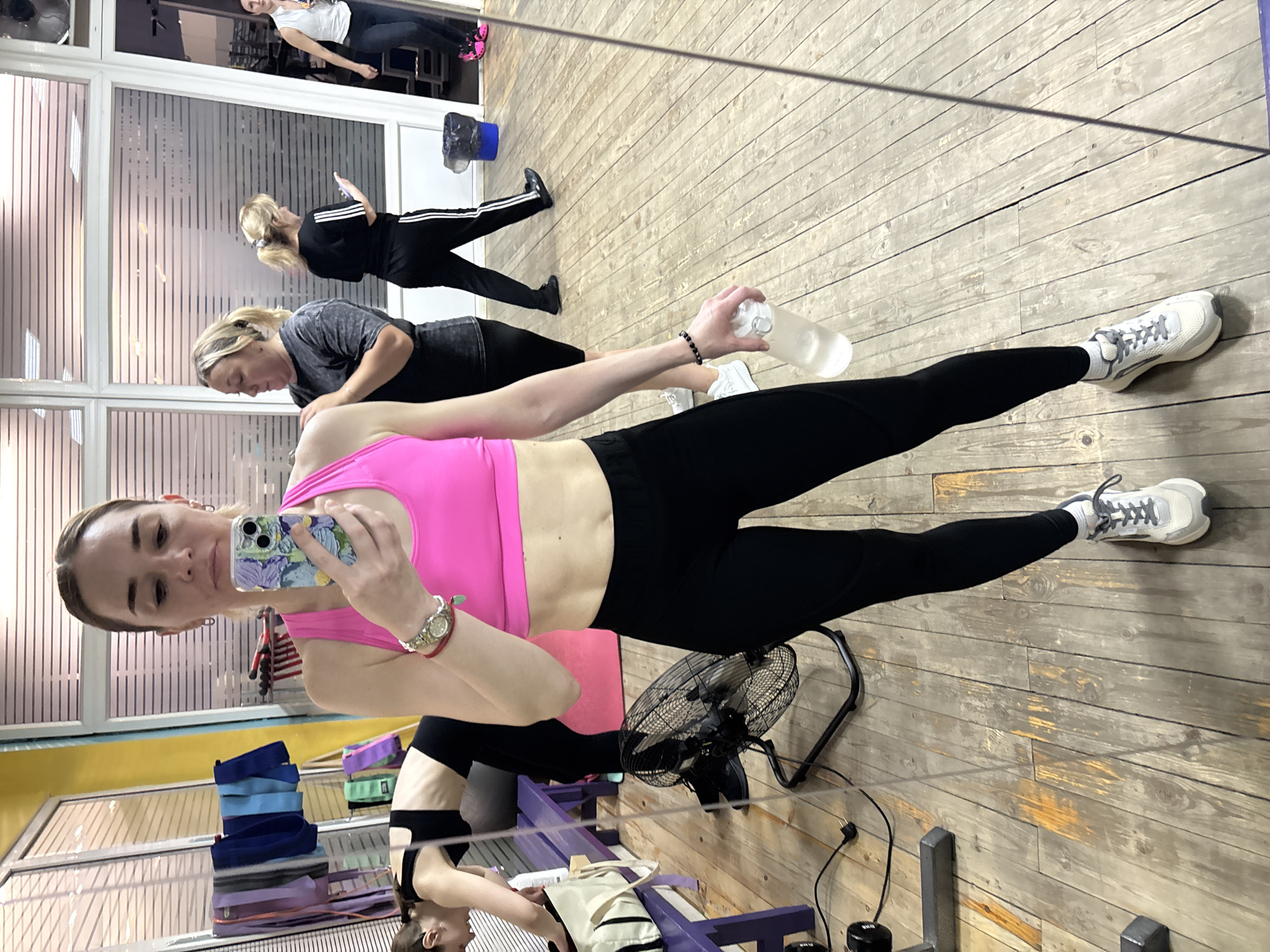A woman is taking a selfie in a gym in Saint Petersburg, Russia. She is wearing a pink sports bra, black leggings, and white athletic shoes. She is holding a clear water bottle in her right hand and her phone in her left. In the background, other women are exercising and preparing for their workouts. The gym has wooden floors and a large mirror reflecting the scene. Equipment such as resistance bands, a fan, and weights are visible. The lighting suggests it is daytime.