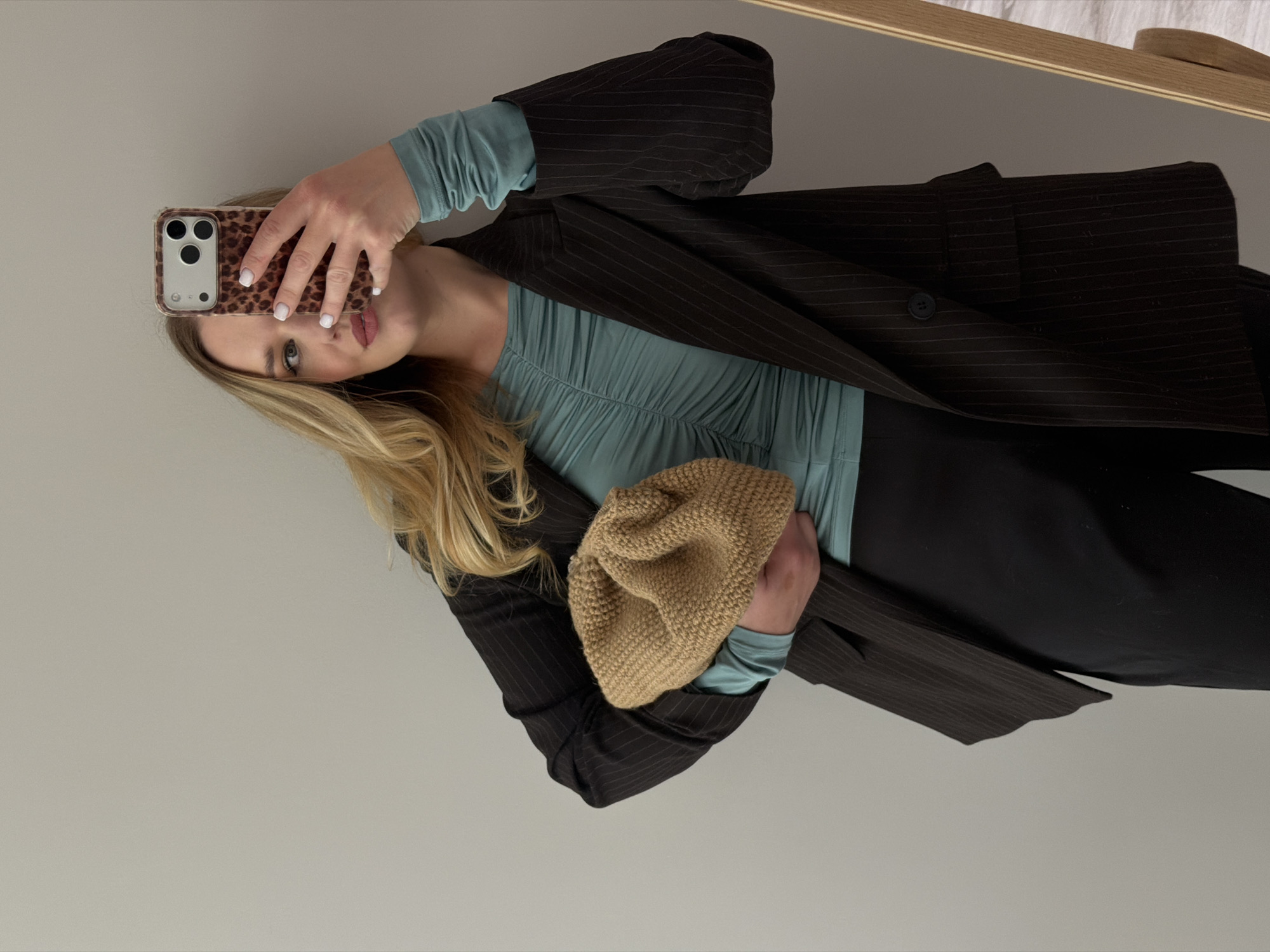 A woman is taking a selfie in a mirror. She is wearing a dark pinstripe blazer over a teal ruched top and black pants. She is holding a tan knitted clutch bag. Her hair is blonde and wavy, and she is wearing light makeup. The background is a plain light-colored wall. The location is Dreiliņi, Latvia.