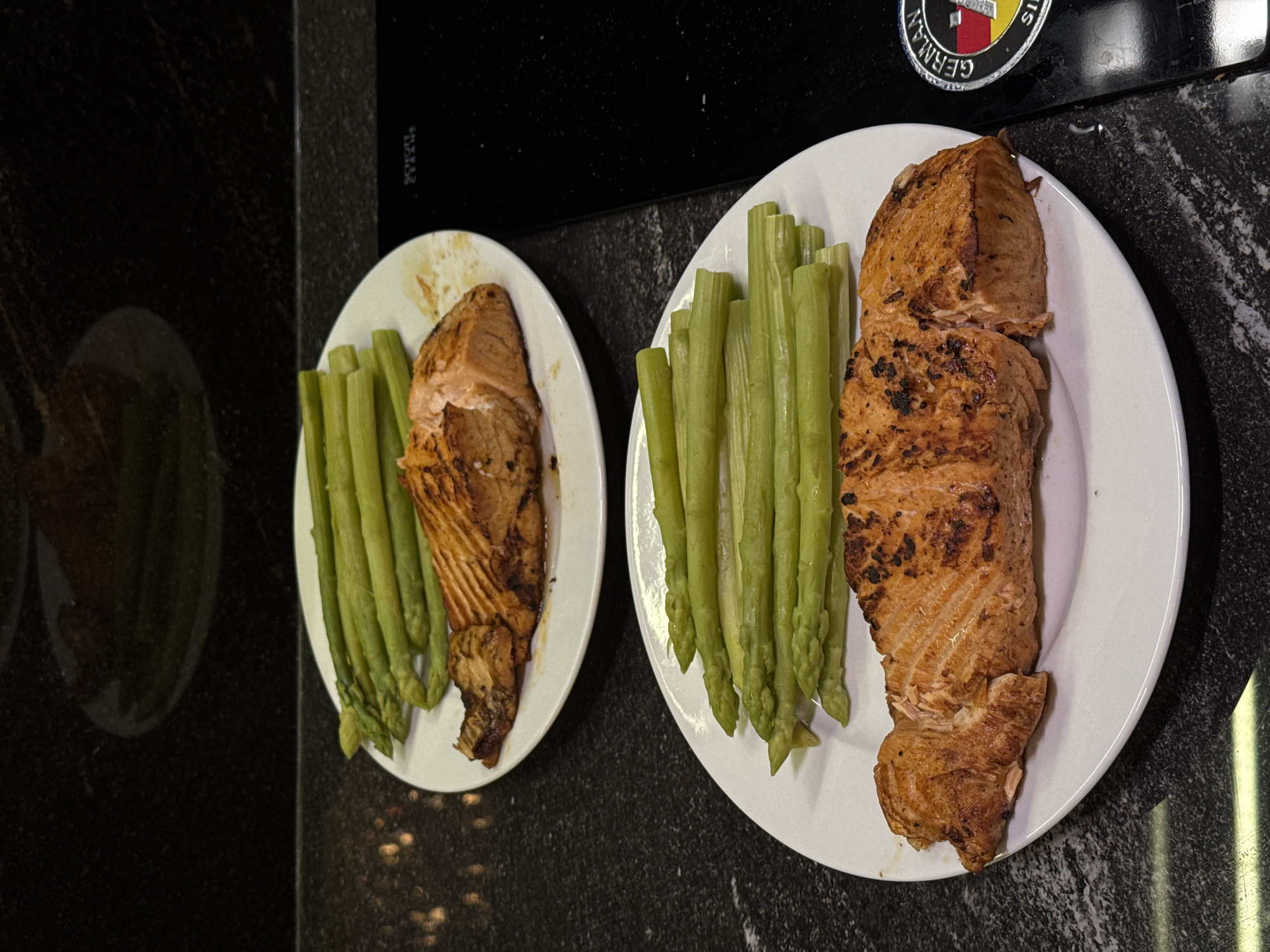 The image shows two white plates, each containing a piece of cooked salmon and a bundle of green asparagus spears. The plates are positioned on a black countertop with a dark, speckled pattern. The lighting appears to be from an artificial source, creating highlights on the food and countertop. The setting seems to be indoors, possibly a kitchen, but the city or location is not identifiable from the image.