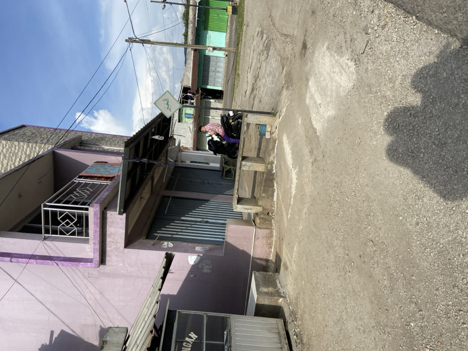 The scene is a street in Sukatani, Indonesia, located at coordinates -6.638 latitude and 107.407 longitude. A man in a pink and purple shirt is seated on a motorbike parked in front of a building with a canopy. The man is facing to the right. The building has a closed, corrugated metal door. There are steps and a bench in front of the building. The street is a gravel surface. There are buildings in the background, including a two-story structure with a green roof. There is a sign with Arabic writing hanging from a pole above the motorbike. There are electrical wires strung overhead. The sky is blue with some clouds.