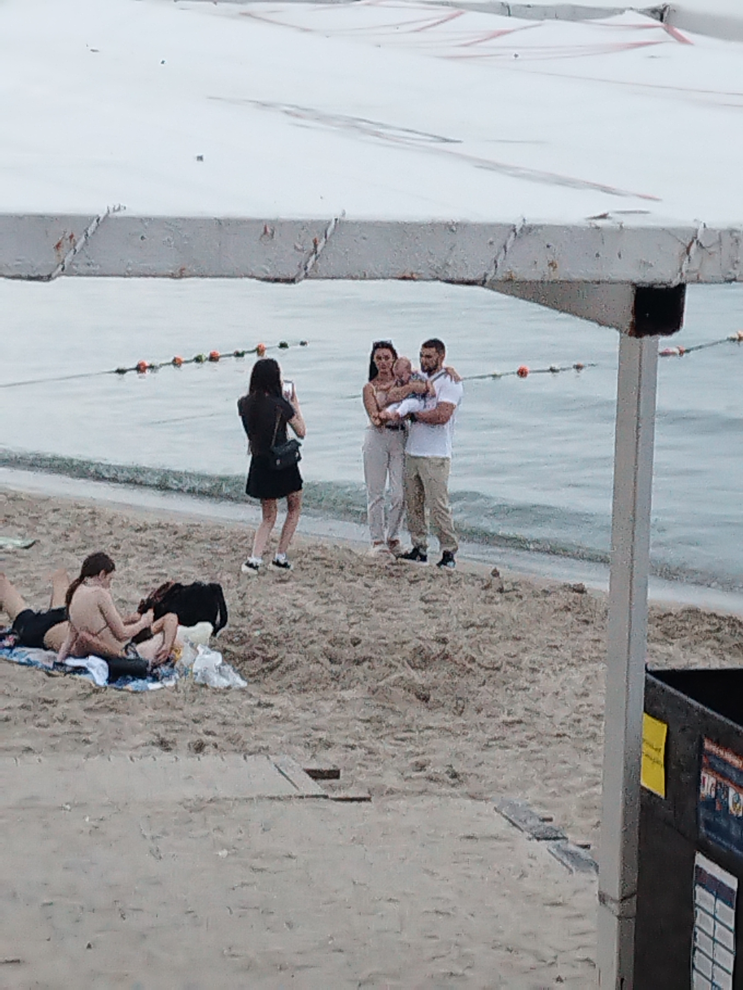 The image shows a beach scene in Odesa, Ukraine. In the foreground, two individuals are lying on a towel on the sand, with one appearing to be tending to the other. Further back, on the shoreline, a woman is taking a photo with her phone of a couple holding a baby. The couple consists of a man and a woman, with the man holding the baby in his arms. The sea is visible in the background, with buoys marking a boundary in the water. A white awning structure is partially visible in the upper portion of the image. The lighting suggests it is daytime, possibly overcast.