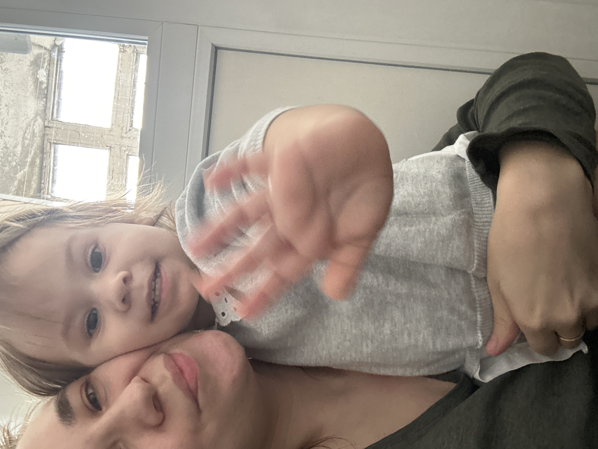 A mother and her young daughter are taking a selfie together indoors. The daughter is leaning on her mother, and she is holding up her hand towards the camera. They are located in Uva, Russia, inside a building with a window visible behind them. The window is multi-paned with grimy glass. The mother is wearing a dark shirt, and the daughter is wearing a light grey sweater with a white collar. The mother has a gold ring on one of her fingers.
