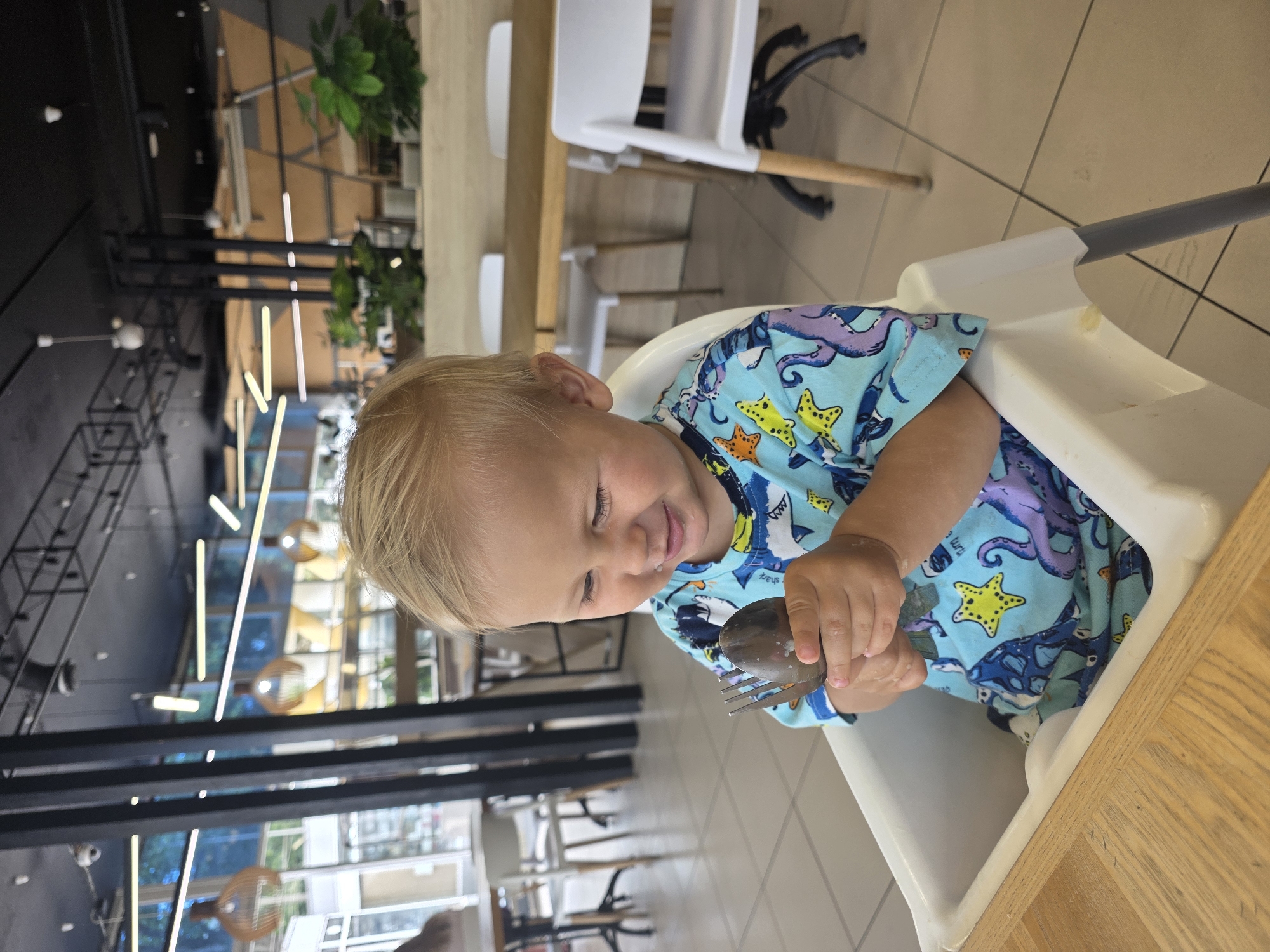 A young child with blond hair is sitting in a high chair in what appears to be a restaurant. The child is wearing a light blue t-shirt with a pattern of sea creatures, including octopuses and starfish. The child is looking down and smiling, holding a fork and spoon in their hands. The high chair is white and is positioned at a wooden table. In the background, there are other tables and chairs, as well as plants and lighting fixtures. The overall scene suggests a casual dining environment.