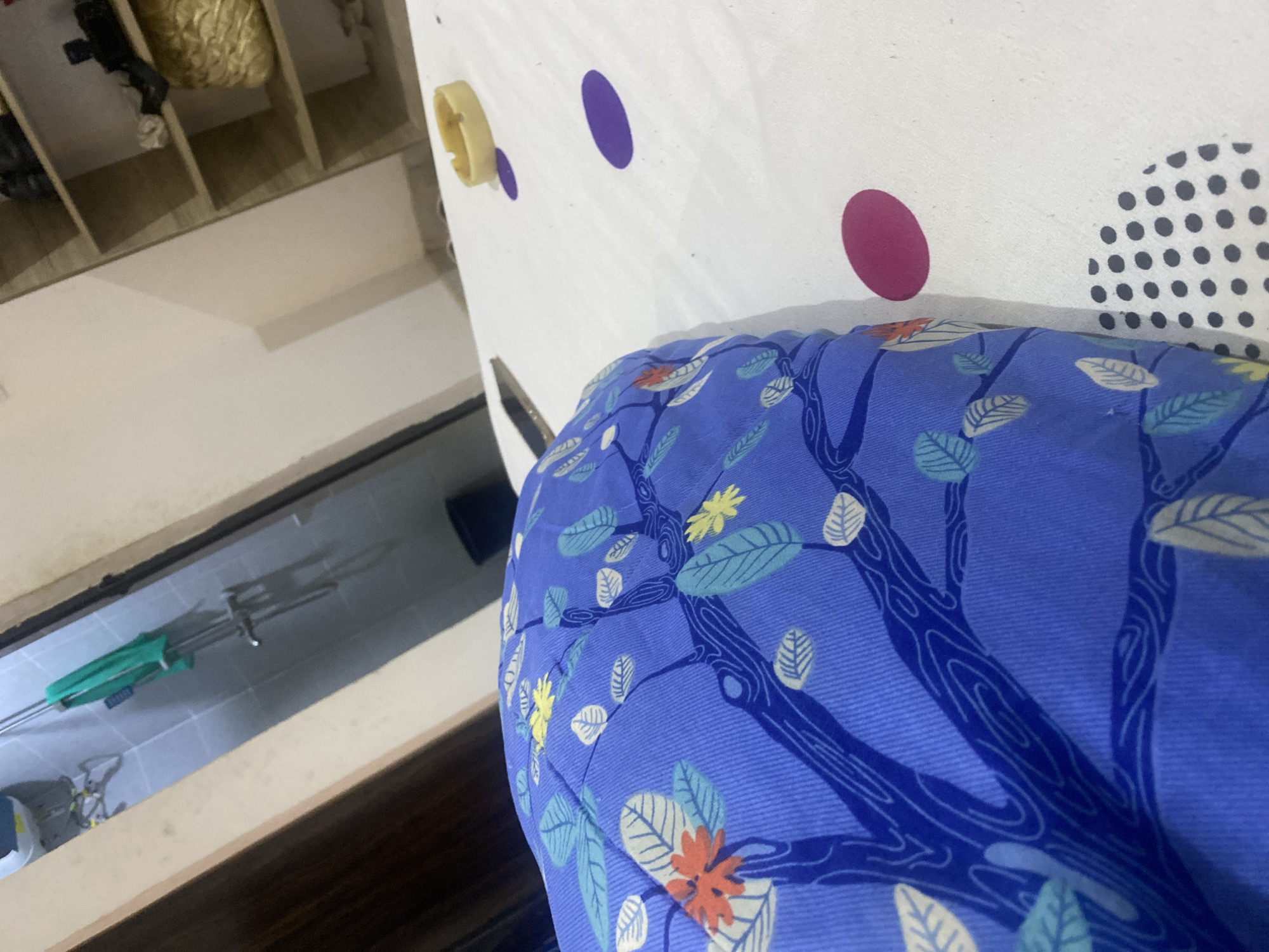 The image presents an indoor scene, prominently featuring a blue floral patterned cushion placed on a surface with multicolored polka dots, next to a shelving unit filled with items. A glimpse of a tiled floor with a metal rack is also visible in the background. The image was taken indoors, the lighting conditions suggest daytime. There are no people visible in the image. The location in Abuja, Nigeria cannot be determined from the visual information provided.