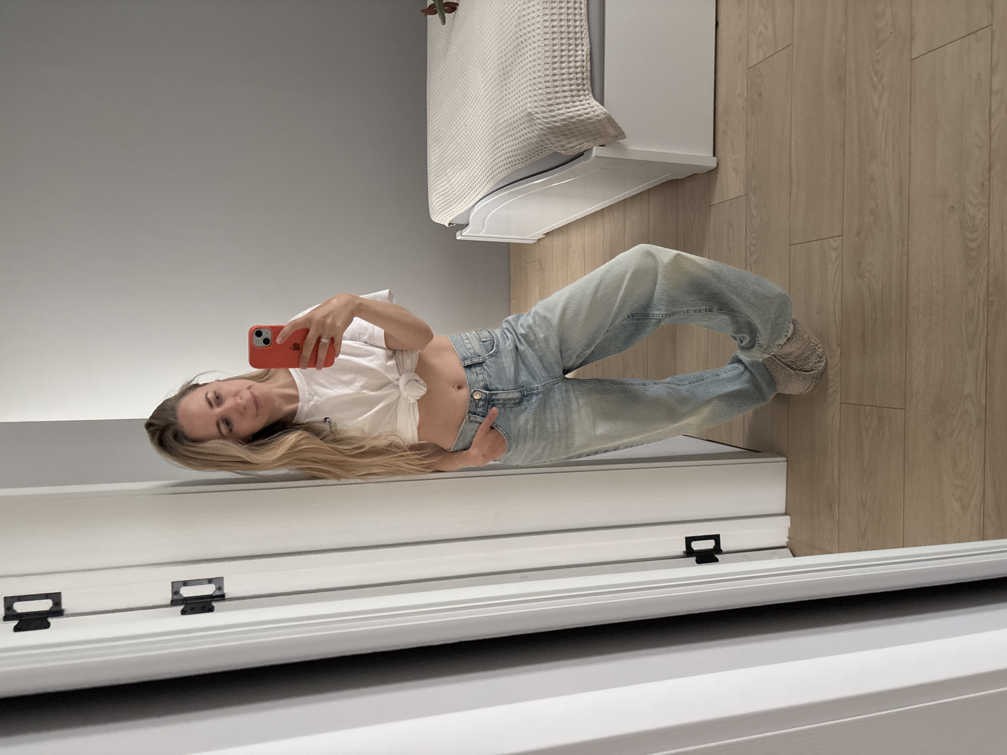 A blonde woman is taking a mirror selfie in a bedroom. She is wearing a white knotted t-shirt and light-wash baggy jeans. The woman is holding a red smartphone in her right hand and has her left hand in her pocket. She is wearing a ring on her left ring finger. In the background, there is a white bed with a textured beige comforter and a light gray wall. To the left of the woman, there is a white door frame with black door hinges. The floor is made of light-colored wood planks. The scene is likely indoors, in a residential setting. Without further information, the location in the city of Chaikovsky, Russia cannot be determined.