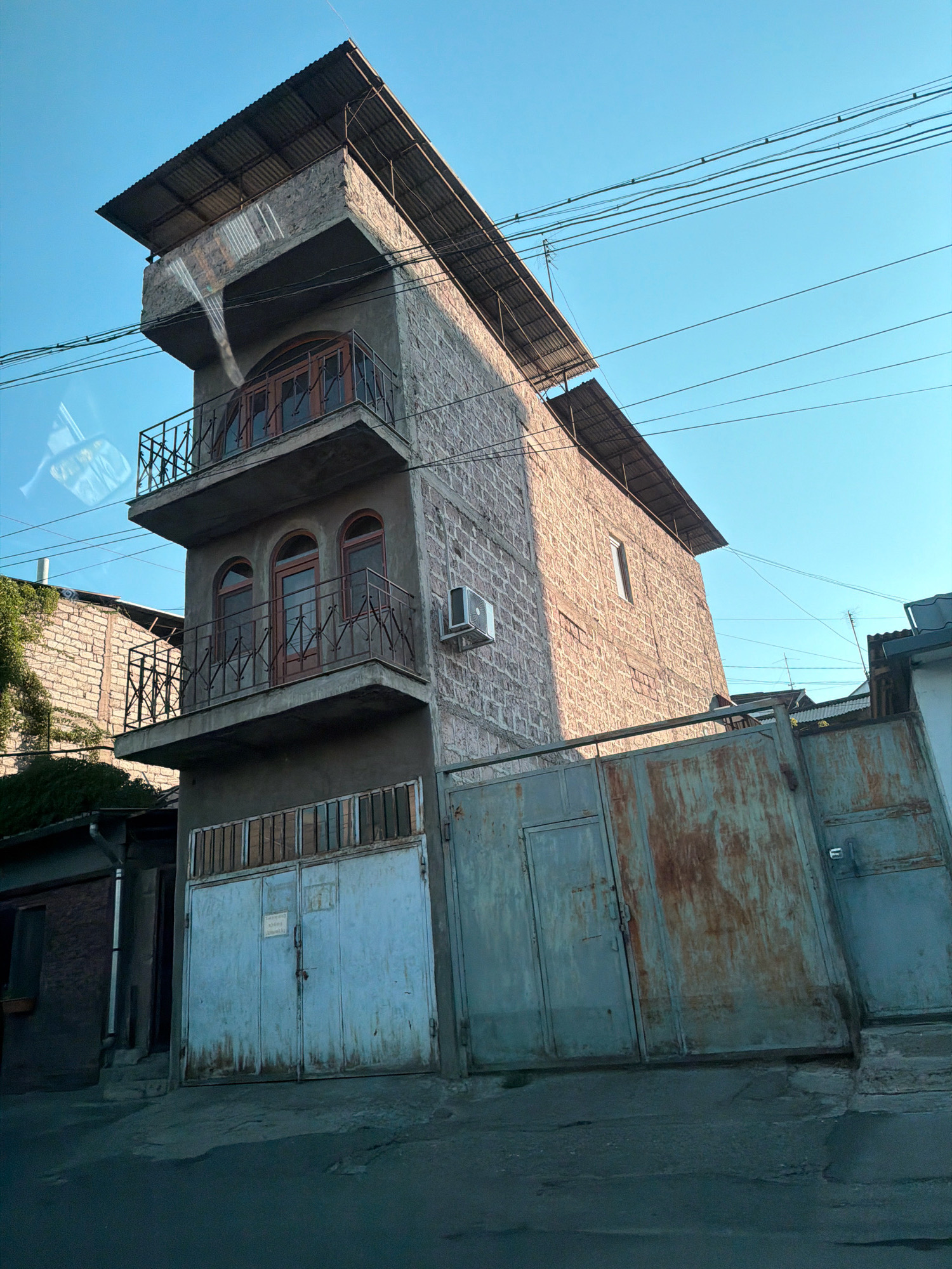 The image shows a narrow, three-story building with a corrugated metal roof. The building is constructed of stone and concrete. It has balconies with wrought-iron railings on the first and second floors. The windows on the second floor are arched and made of wood. An air conditioning unit is mounted on the exterior wall of the building. In front of the building are two large, rusty metal gates. A blue-gray, multi-paneled gate is on the left, and a larger, more weathered gate with rust streaks is on the right. Overhead, a tangle of power lines crisscrosses the clear blue sky. The street in front of the building is asphalt. The building appears to be in a residential area of Yerevan, Armenia, judging by the architectural style and the presence of overhead wires.