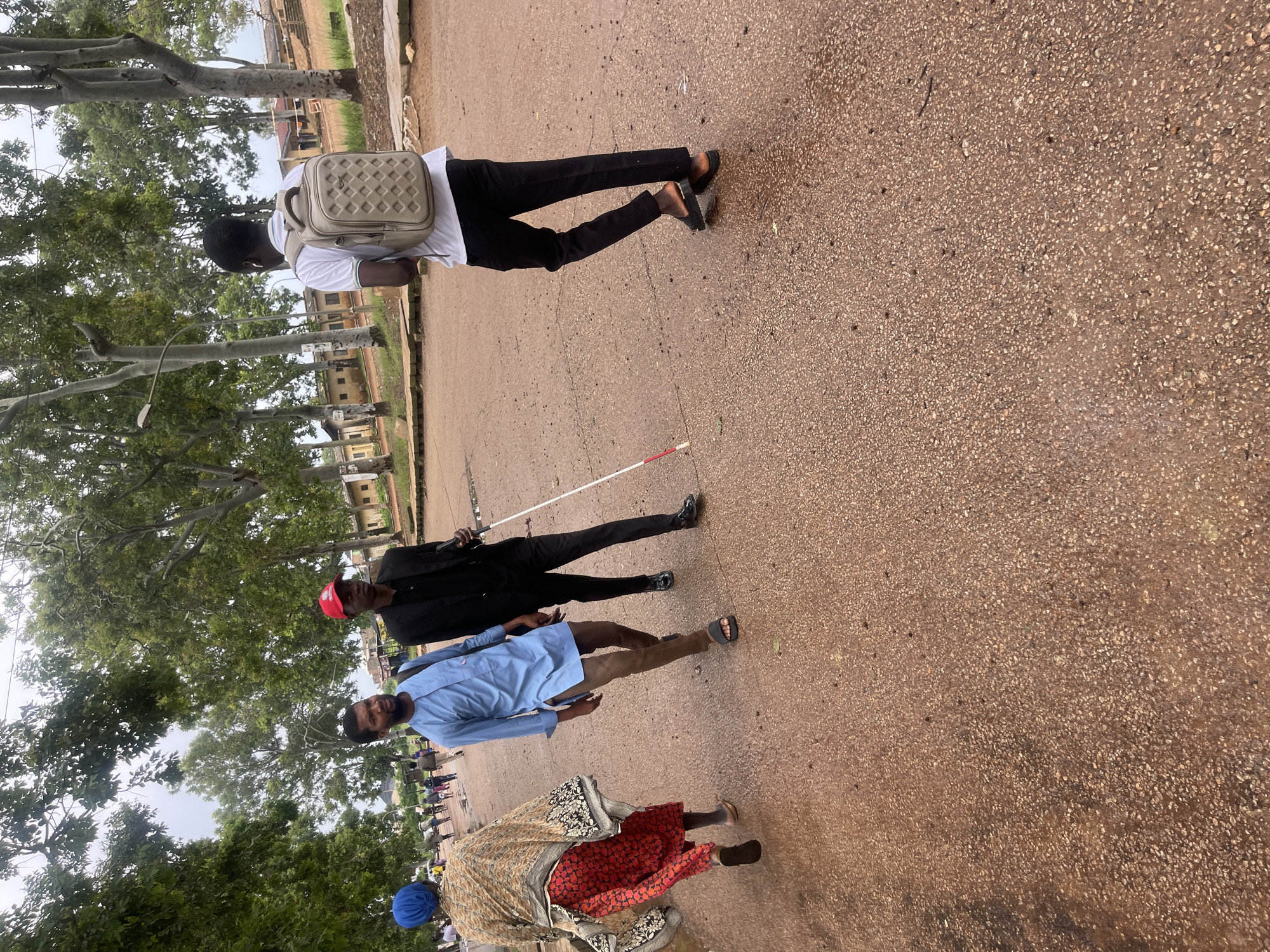 The image captures an outdoor scene in the city of Rafin Par, Nigeria, likely on a cloudy day given the diffused lighting. The ground is a reddish-brown asphalt or dirt road with some wet patches. In the foreground and mid-ground, four individuals are walking.
On the far left, a woman wearing a blue headscarf and a colorful patterned wrap over a red skirt walks away from the camera.
To her right, two men are walking together. The man on the left is wearing a light blue traditional African tunic and brown trousers, with sandals on his feet. He is looking towards the left. The man on the right is wearing a black suit and a red cap, and he is holding a white cane with a red tip, indicating he is visually impaired. He is using the cane to navigate.
Further to the right and slightly behind the two men, a man wearing a white polo shirt and black trousers walks away from the camera, carrying a light beige backpack with a patterned front.
In the background, there are trees with green foliage and a few low-rise buildings with reddish-brown roofs. Other people are visible walking in the distance, suggesting this is a public area. The overall impression is a casual street scene with people going about their day.
