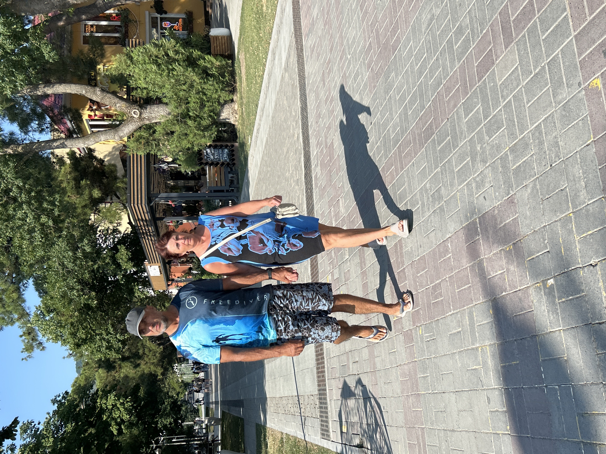 A man and a woman are walking hand-in-hand on a paved walkway in Gelendzhik, Russia. The man is wearing a blue t-shirt with a graphic print and patterned shorts. The woman is wearing a blue and black floral dress and carrying a small white purse. They are walking towards the viewer's right. The walkway is lined with trees and buildings. The sky is clear and blue.