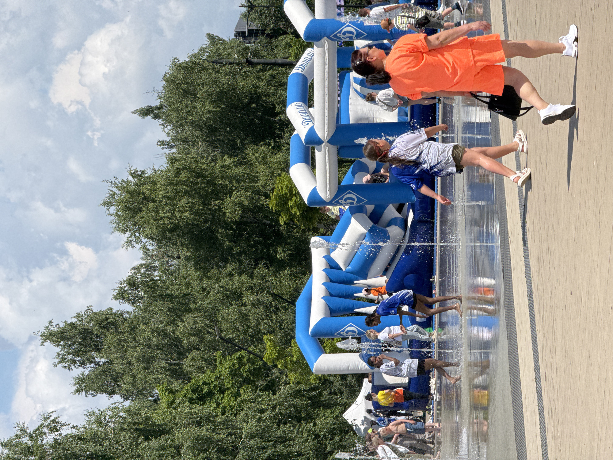 On a sunny day, a group of people are enjoying a water park or splash pad area in Izhevsk, Russia. In the background, a large blue and white inflatable structure with the word "Динамо" (Dynamo) printed on it, possibly a bouncy castle or obstacle course, is set up. Water jets spray from the ground in the foreground, creating a shallow pool where several children and adults are playing.
In the mid-ground, a young girl in a white and blue shirt with olive shorts walks through the water, facing away from the camera. To her right, another young person wearing a blue shirt and dark shorts is also in the water, looking towards the inflatable structure. Behind them, several other individuals, including a child in an orange shirt, are visible.
In the foreground, a woman wearing a bright orange t-shirt and matching shorts walks away from the camera, carrying a black bag. She is wearing white sneakers. To her left, a young person in a white and blue t-shirt and olive shorts walks in the same direction.
The scene is set against a backdrop of lush green trees, with a bright blue sky and scattered white clouds overhead. The ground is paved with wooden planks in the foreground, transitioning to a wet, reflective surface where the water feature is located.