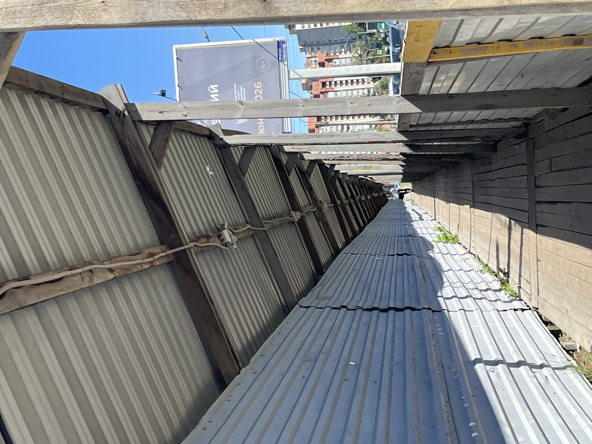 This image depicts an outdoor scene with a focus on a long, corrugated metal structure that appears to be a covered walkway or part of a construction site. The structure is made of two parallel corrugated metal roofs, angled downwards and extending into the distance. The roof on the left is supported by wooden beams and appears to be attached to a corrugated metal wall, also supported by wooden framing. Electrical wiring is visible running along one of the wooden support beams on the left side. The roof on the right is also made of corrugated metal and is supported by wooden beams that are part of a wooden wall structure. There are patches of greenery, possibly weeds or small plants, growing between the metal sheets and along the base of the wooden wall on the right.
In the background, beyond the structures, a clear blue sky is visible, suggesting fair weather during the daytime. Several multi-story buildings with brick facades are visible, indicating an urban environment. A large billboard with text is also present in the background, featuring Cyrillic script. The text on the billboard is partially obscured but includes words like "ий" and "026". The overall scene gives the impression of a functional, possibly temporary, structure in a city setting. The lighting suggests it is daytime, likely sunny given the shadows cast by the wooden beams. There are no people visible in the image.