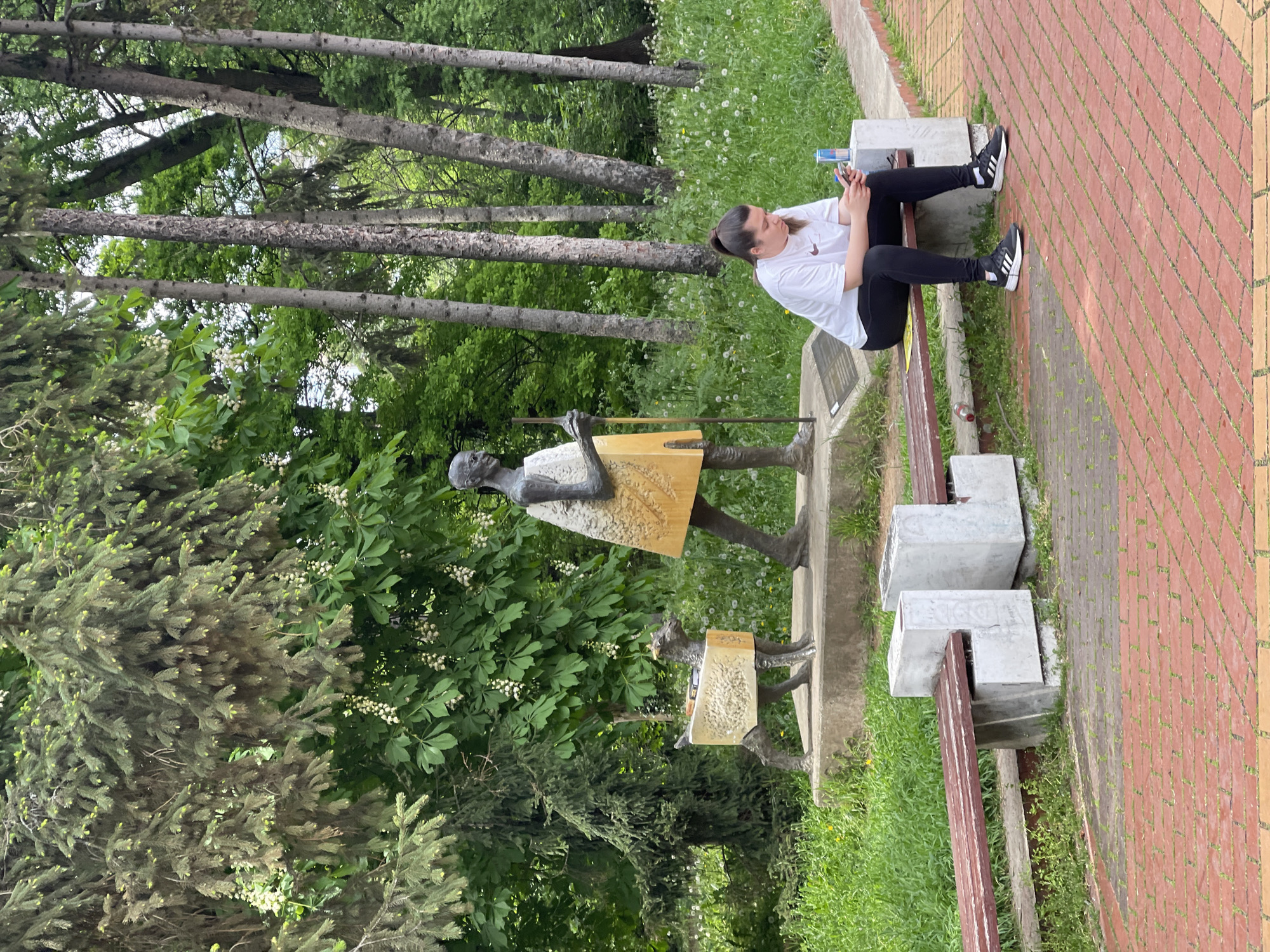 A young woman sits on a park bench, absorbed in her phone, in Sofia, Bulgaria. The bench is located near a statue depicting a group of figures carrying rectangular burdens. The scene includes trees, grass, and a brick pathway.