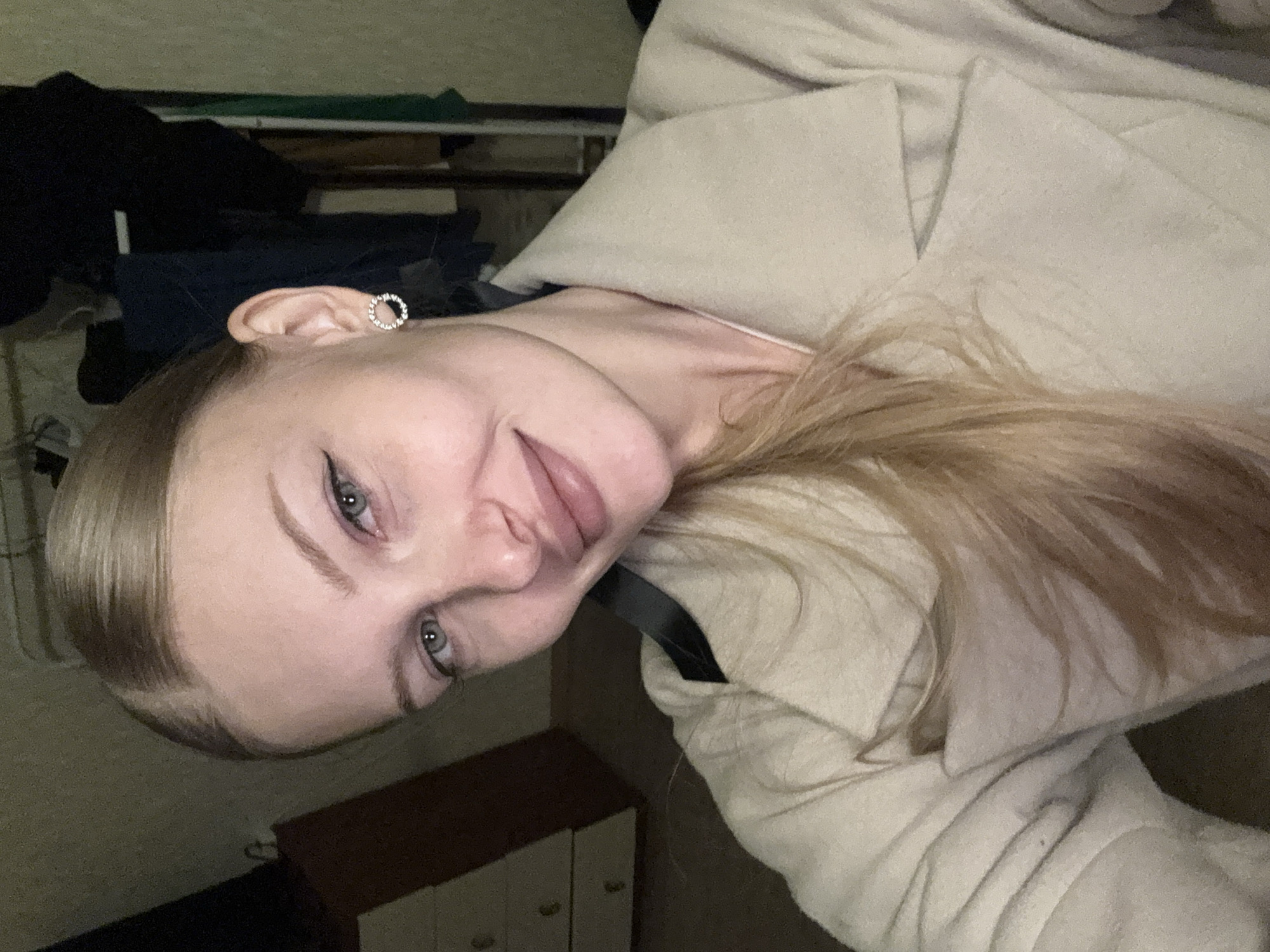 A young woman with blonde hair pulled back into a ponytail is taking a selfie in a dimly lit room. She is wearing a light-colored coat. She has on subtle makeup, including eyeliner and lip gloss, and small, circular earrings with rhinestones. In the background, a dresser and clothing rack are visible. The setting appears to be a domestic interior, potentially in Kondratovo, Russia.