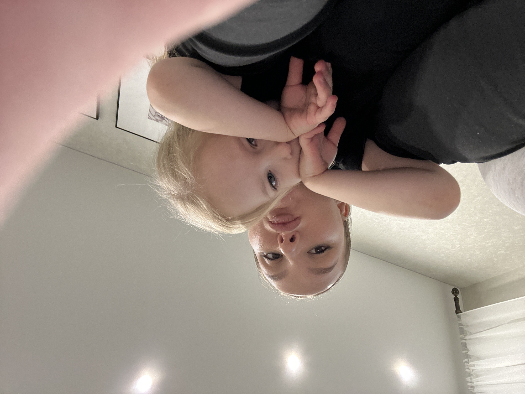 The image shows a Caucasian woman and a young child, likely a toddler, in an indoor setting. The photo is taken from an unusual, low angle, looking upwards. The woman is oriented "upside down" in the frame, with the child positioned above her chest and partially obscuring her face. The child has fair hair and is holding their hands near their face. Both individuals are looking directly at the camera. The woman is wearing what appears to be a dark-colored shirt or dress. The background is a plain, light-colored wall with some indistinct elements suggesting a domestic interior, possibly a bedroom, including curtains and framed artwork. There is no indication of the location being the city of Chaikovsky, Russia or any specific city.