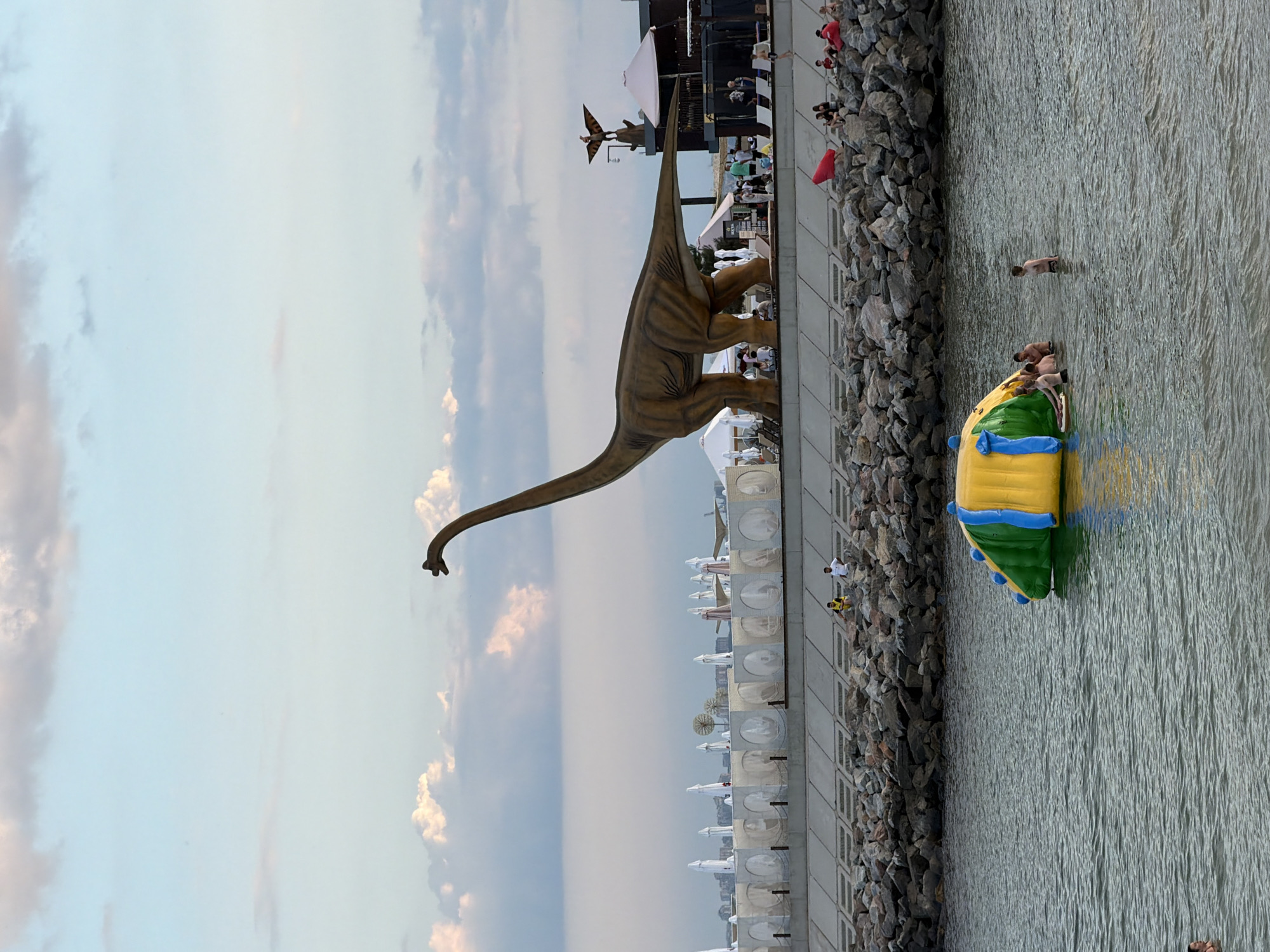 This image captures a waterfront scene, possibly at the Fontanka River embankment in Odessa, Ukraine, given the style of the promenade and the presence of large scale decorative elements. In the foreground, the choppy, greenish water of a body of water is visible, with a colorful inflatable water toy in the center. Several individuals are in the water, some near the inflatable structure, suggesting recreational activity.
Dominating the middle ground is a large, tan-colored statue of a Brontosaurus, a sauropod dinosaur, standing on a paved promenade. The promenade is lined with large rocks and concrete barriers, forming a seawall or embankment. Behind the dinosaur, a low-rise building complex is visible, featuring white umbrellas and what appear to be covered seating areas, indicative of a beachside establishment or resort. A smaller decorative statue of a winged creature atop a lamppost is also noticeable on this structure.
The background is a soft blue sky with scattered, fluffy white and gray clouds, suggesting late afternoon or early evening. The overall atmosphere appears to be one of a leisure area, likely a tourist attraction or a popular spot for recreation, with people enjoying the water and the unique dinosaur sculpture.
