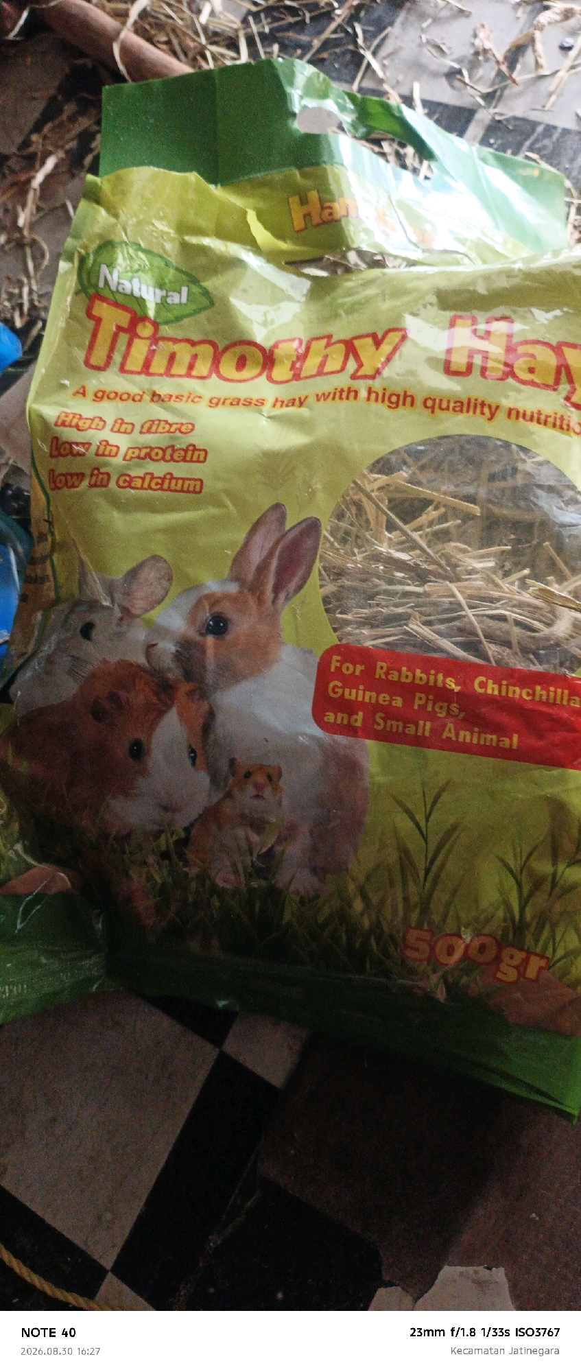 A close-up photograph shows a bag of Timothy hay, a type of animal feed. The bag is predominantly yellow with green accents and features images of rabbits and guinea pigs. Text on the bag indicates it is suitable for rabbits, chinchillas, guinea pigs, and other small animals, and highlights "high fibre," "low in protein," and "low in calcium." A portion of the bag is transparent, revealing the hay inside. The bottom right corner of the image displays camera metadata: "23mm f/1.8 1/33s ISO3767" and "Kecamatan Jatinegara." This indicates the location as Jatinegara District, East Jakarta, Indonesia. The bag rests on a surface with a black and white checkered pattern, likely a floor. The overall lighting is subdued, suggesting an indoor or shaded environment.