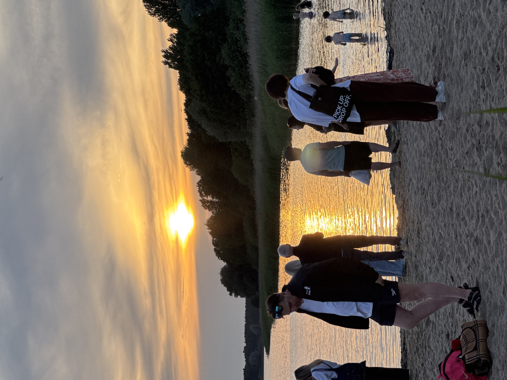 The image depicts a group of people on a sandy beach next to a body of water at sunset. The sky is filled with dramatic clouds illuminated by the setting sun, casting a golden and orange glow on the water. In the foreground, several individuals are standing on the sand, some appearing to be conversing or observing the sunset. Further back, a few people are wading in the shallow water. The background shows a line of trees and reeds along the water's edge. The overall atmosphere is serene and evokes a sense of a relaxed evening. This scene is likely located at a beach in the city of Tallinn, Estonia, given the commonality of such picturesque sunset views over coastal areas or lakes within or near the city.