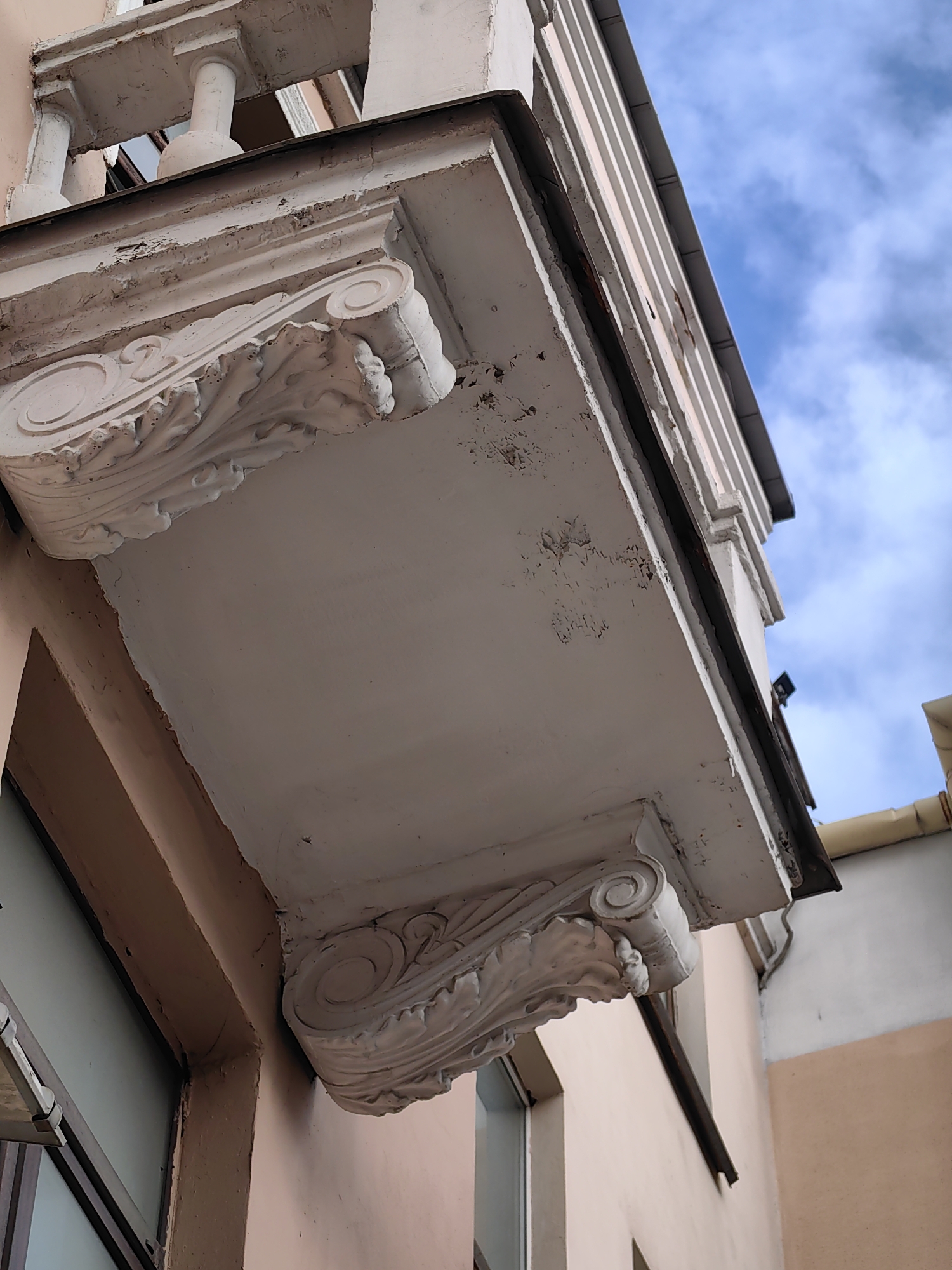 This image captures a low-angle view of a weathered, ornate balcony structure on a building, against a backdrop of a cloudy blue sky. The balcony is supported by decorative corbels featuring scrolled volutes and acanthus leaf motifs, all rendered in white stucco. The white paint on the balcony and its supports is peeling and chipped in several areas, revealing the underlying material and suggesting age and exposure to the elements. The building wall beneath the balcony is painted a pale pinkish-peach color, and a portion of a window with a metal frame is visible on the left. The top edge of the balcony shows a dark metal trim, and to the right, a portion of a light-colored gutter is visible against the building's facade. The overall impression is one of architectural detail in a state of disrepair, set against an outdoor urban environment. The provided crops highlight specific areas of the corbels, peeling paint, and the sky, offering a closer look at the texture and condition of the balcony. No people or specific actions are depicted. Based on external information, this scene is located in the city of Minsk, Belarus.