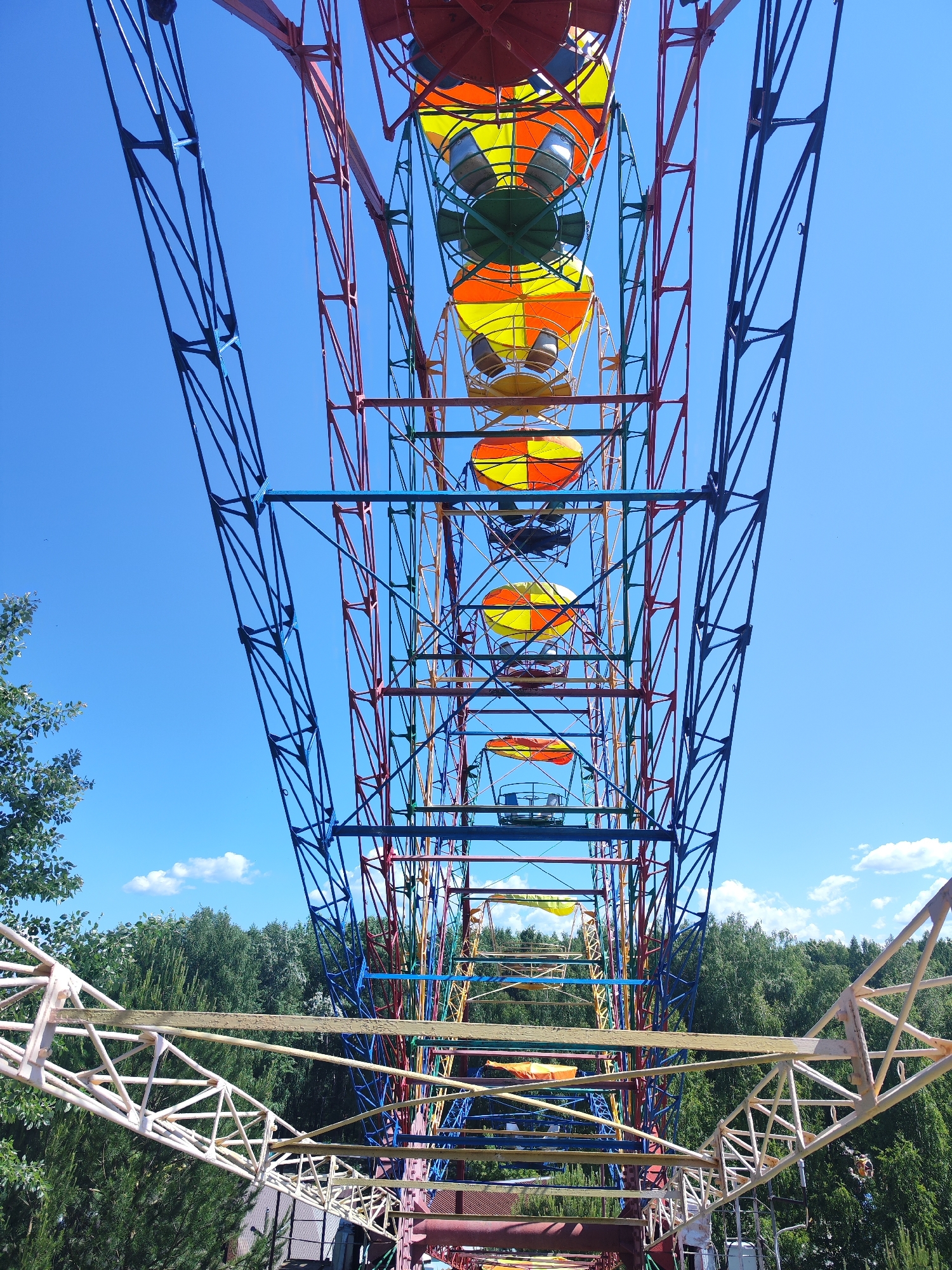 This is a low-angle, upward-looking shot of a Ferris wheel against a bright blue sky with scattered white clouds. The Ferris wheel is constructed of brightly colored metal struts, including blue, red, yellow, and green. The gondolas, also colorful with yellow and orange patterns, are visible at various points along the wheel's circumference. The structure of the Ferris wheel dominates the frame, extending upwards and towards the viewer. In the background, a dense green forest canopy is visible, suggesting an outdoor setting. On the lower left, a portion of a white metal support structure for the Ferris wheel is visible, along with some tree branches and foliage. The image appears to have been taken during daylight, with strong shadows indicating a sunny day. The context suggests this Ferris wheel is located in an amusement park or recreational area, possibly in the city of Chaikovsky, Russia, given the prompt's request. There are no people or animals clearly visible in the frame.