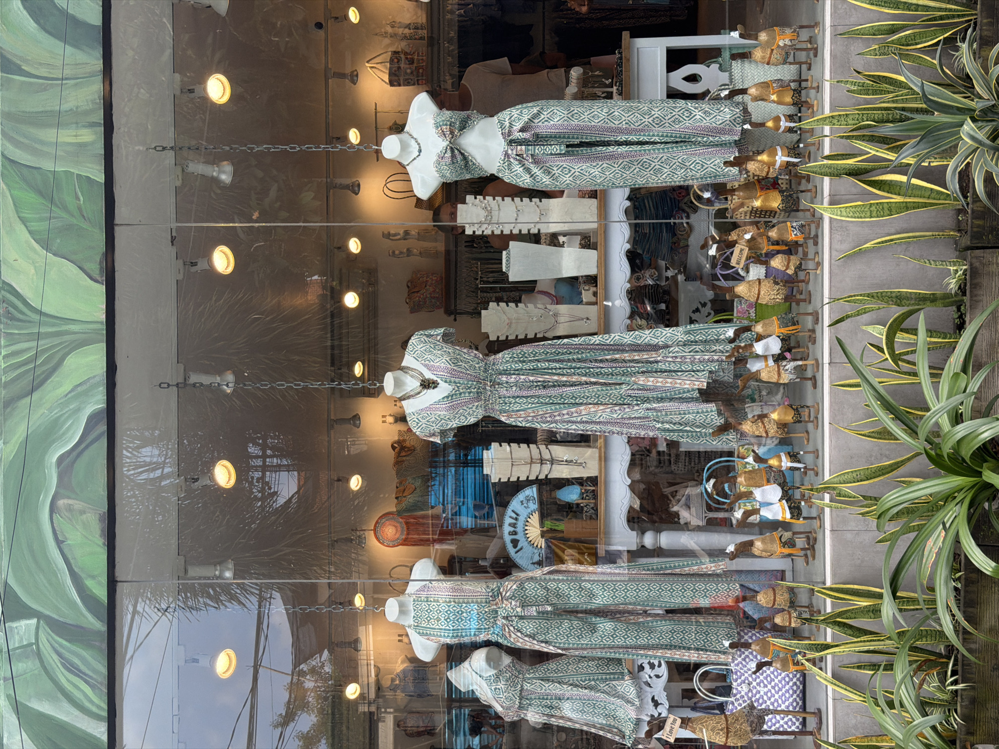 This is a full, eye-level, outdoor shot of a store window display in Ubud, Indonesia, featuring apparel and decorative items. The window has a large mural of green leaves painted across the top. Inside, mannequins are dressed in patterned jumpsuits and dresses, with various necklaces displayed around them. A white table displays more jewelry and a fan that says "I LOVE BALI." The foreground is filled with potted plants with long, green and yellow striped leaves. In front of the window, a collection of wooden duck figurines, some with hats and clothing, are arranged. The lighting appears to be artificial, with spotlights illuminating the store's interior. Reflections of the sky and trees are visible in the glass.