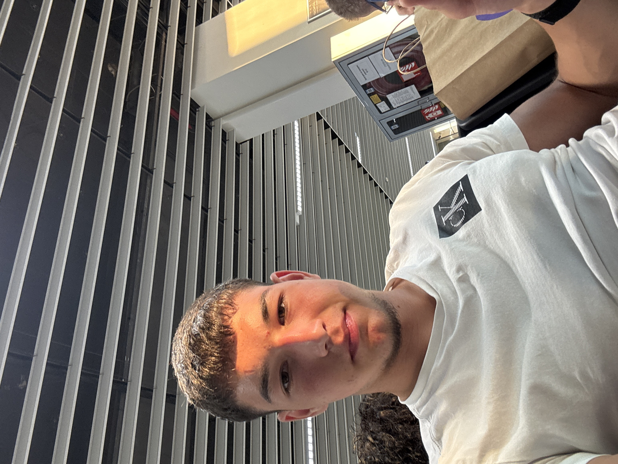 The image shows a close-up, rotated view of a young man from the chest up. He is wearing a white t-shirt with a black logo on the left breast. The logo appears to be a stylized "CK" within a square or diamond shape. The man has short, dark hair and a light beard. Sunlight is casting a warm glow across the left side of his face, highlighting his features. He has a slight smile and appears relaxed.
Behind him, there are vertical metallic slats, suggesting an indoor or covered outdoor space with a modern architectural design. In the upper right of the image, a brown paper bag with its handle visible is held by someone out of frame, and to its left, a display board with some text and graphics is visible. The lighting suggests it is daytime, possibly late afternoon due to the warm, angled sunlight. The overall scene seems to be a public area, perhaps a waiting area or a casual dining establishment, given the presence of the paper bag and display board. The location context provided is Sofia, Bulgaria.