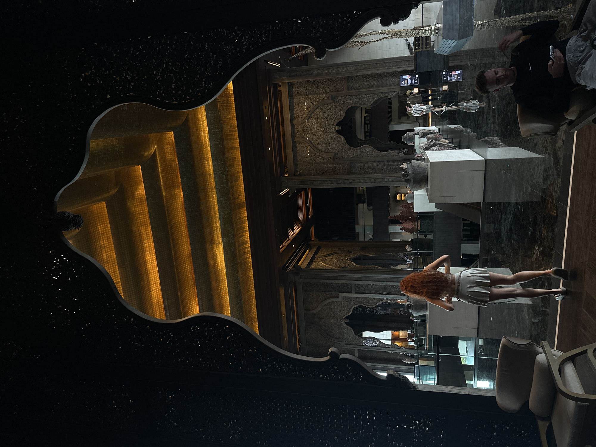 Captured in Nusa Dua, Indonesia, the image presents a reflected scene within a decorative, dark-toned frame. Inside the frame, illuminated vertical stripes of warm light contrast with the dark interior. The reflection encompasses a modern interior, including a woman in a light-colored dress and a man seated with a mobile phone. Art installations and a contemporary design are visible in the mirrored space. The environment suggests a luxury hotel or upscale resort lobby.