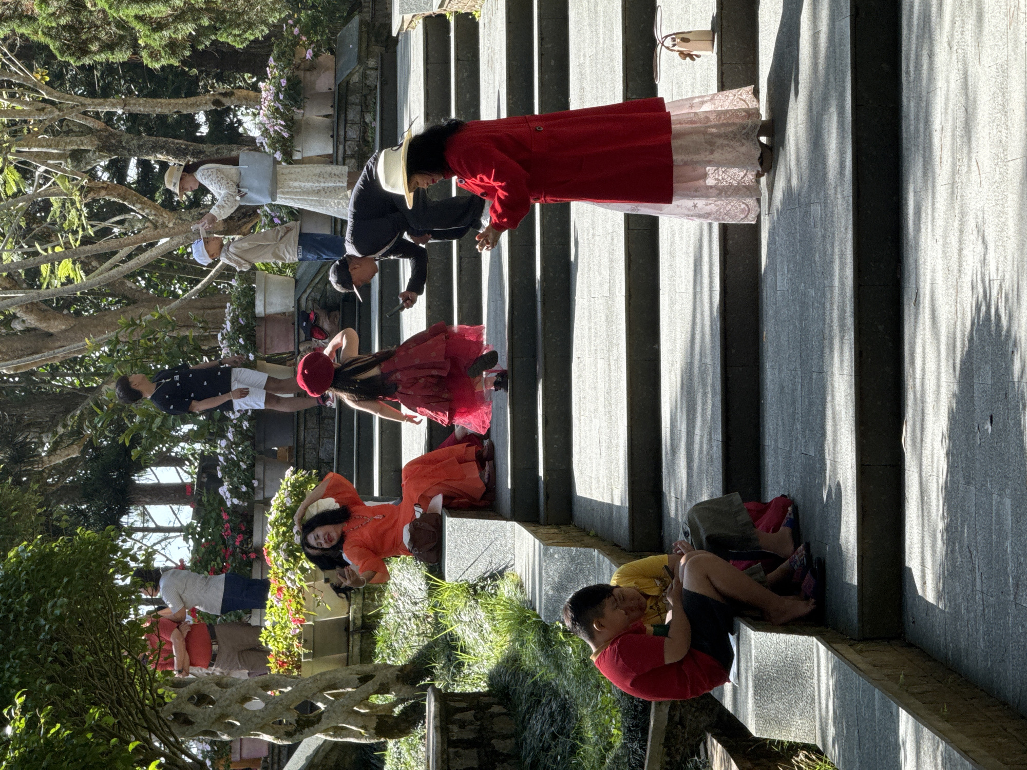 In Da Lat, Vietnam, on a sunny day at a botanical garden or park, several people are seen on a stone staircase and surrounding area. In the foreground, two young boys are seated on a stone ledge, one in a red shirt and the other in a yellow shirt, both looking at a mobile phone. Further up the stairs, a woman in an orange dress is taking a selfie with her phone, with a brown bag resting beside her. A young girl in a red dress and a red hat is walking up the stairs. To her left, a man in a dark suit and cap is also on the stairs, holding a phone and appearing to take a photo. On a higher step, a woman in a white dress and a straw hat is holding a phone, and a young boy in a beige hoodie and white cap stands near her. Further up, a man in a red shirt and brown pants is talking to a woman in a grey shirt and blue jeans. The background is filled with trees and potted flowers. The atmosphere suggests a casual outing or visit to a tourist attraction.