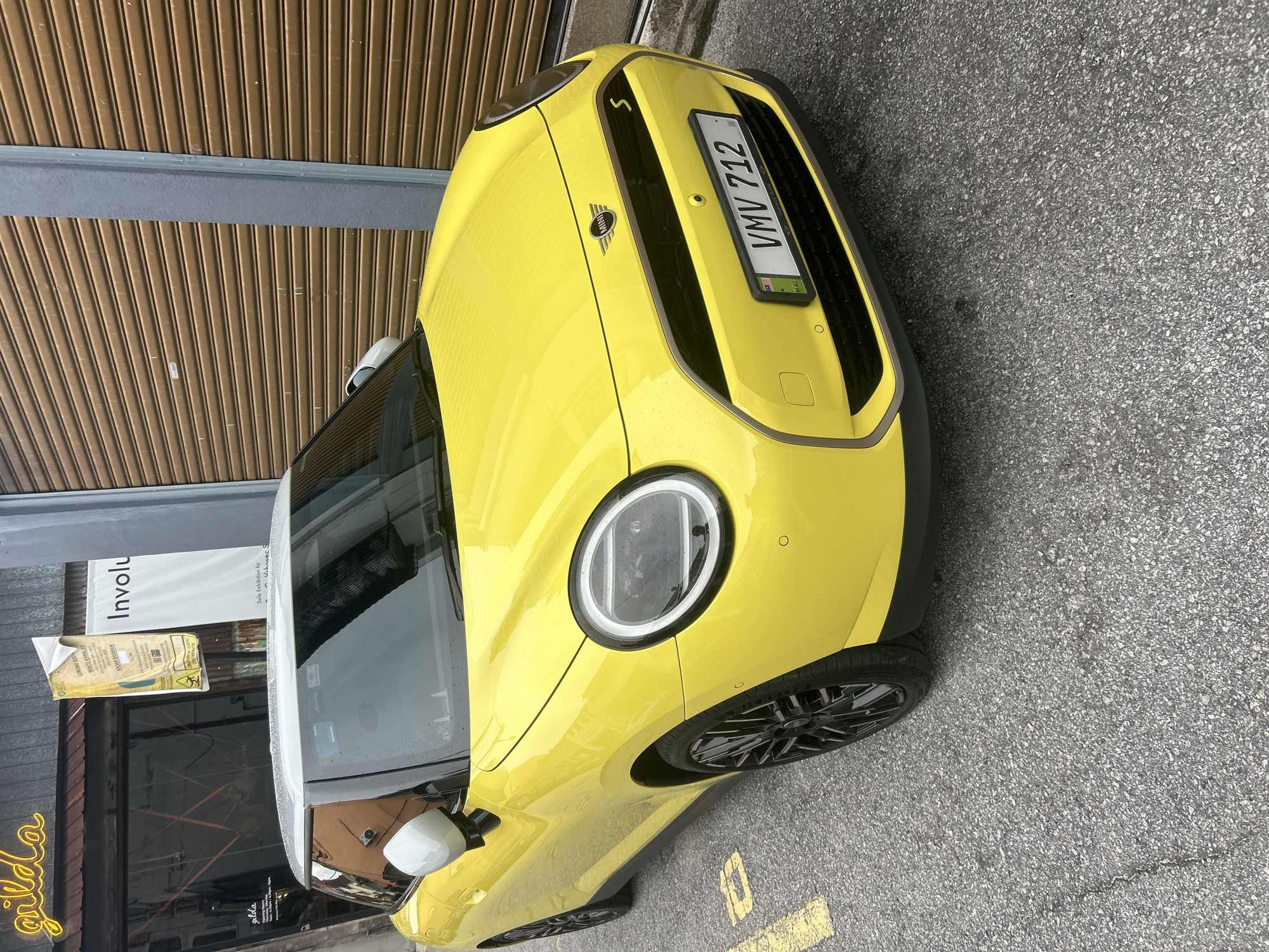 A bright yellow Mini Cooper with a white roof is parked on a paved street in George Town, Malaysia. The car is facing slightly to the right, and its license plate reads "VMV 712". In the background, there is a building with a large, rolling metal shutter and signage that includes the word "Involu". Another sign above the entrance to the building reads "gilda" in a neon script. The street is marked with yellow lines. The weather appears to be overcast, and there are visible raindrops on the car's surface, suggesting it might be raining or has recently rained. The building's facade also shows water stains and some posters. The overall scene depicts an urban streetscape with a prominent yellow car as the central subject.