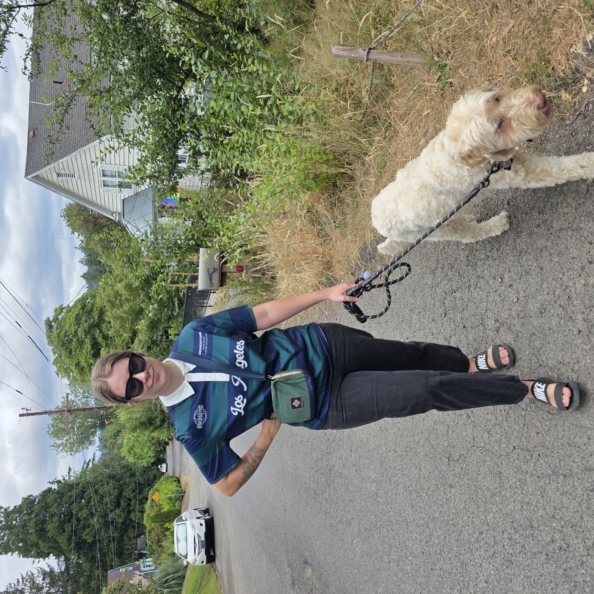 In Portland, Oregon, on a cloudy day, a young woman with blonde hair and sunglasses walks her light-colored, curly-haired dog on a leash down a paved residential street. She is wearing a dark blue and green striped collared shirt with "Los Angeles" written across it, black pants, and black Nike slides. A small green crossbody bag is worn over her shoulder. The dog, which appears to be a Doodle breed, is walking ahead of her on the right side of the street. The street is lined with trees and houses, one of which is visible on the right side of the frame. The environment suggests a suburban setting with overgrown dry grass and foliage along the side of the road.