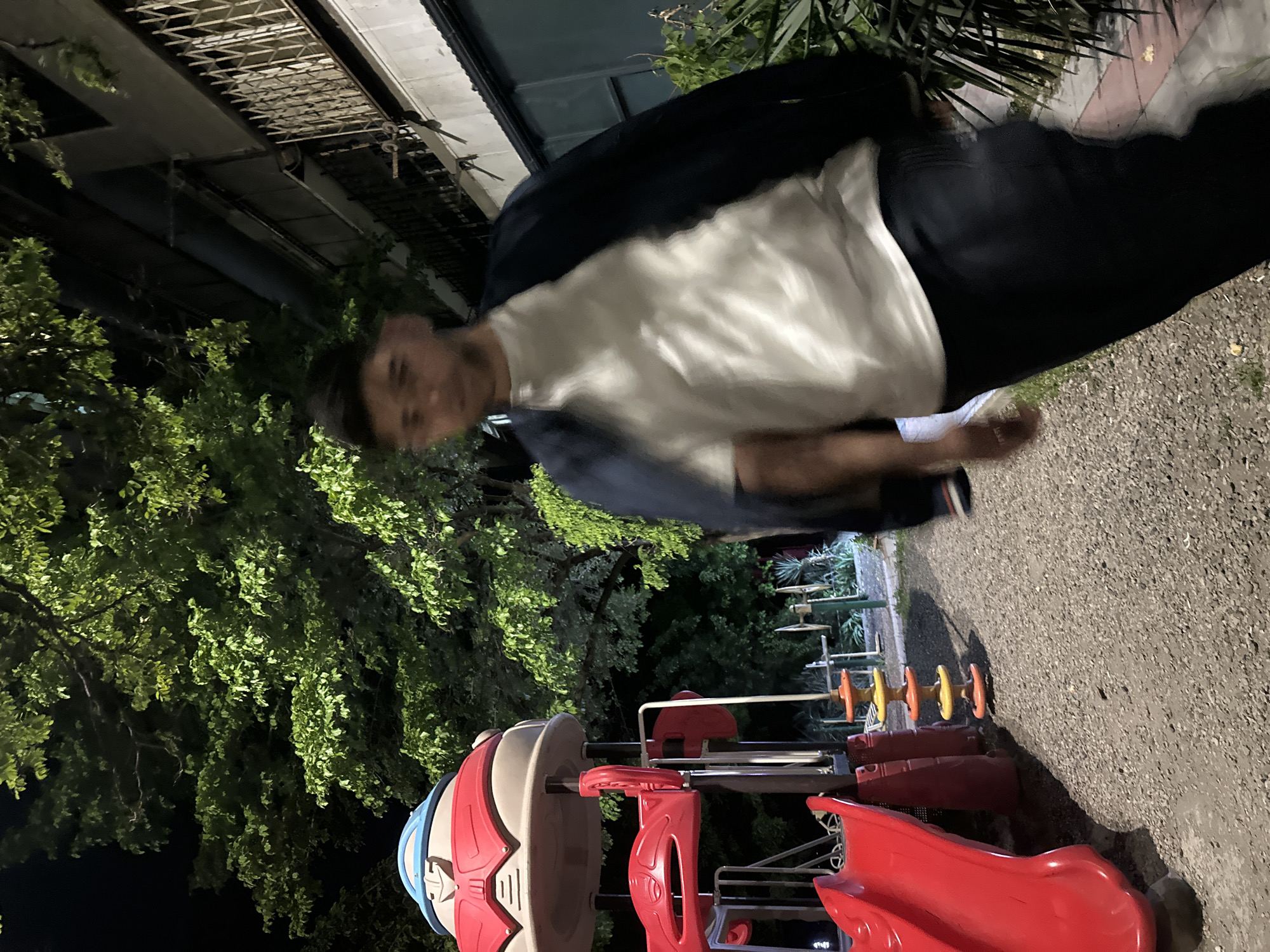 The image shows a man wearing a white t-shirt and dark jacket, standing in what appears to be a playground. A red and white playground structure is in the foreground. Trees surround the playground, and a building is visible in the background. The ground is covered in gravel. Yerevan, Armenia is not discernible from the image.