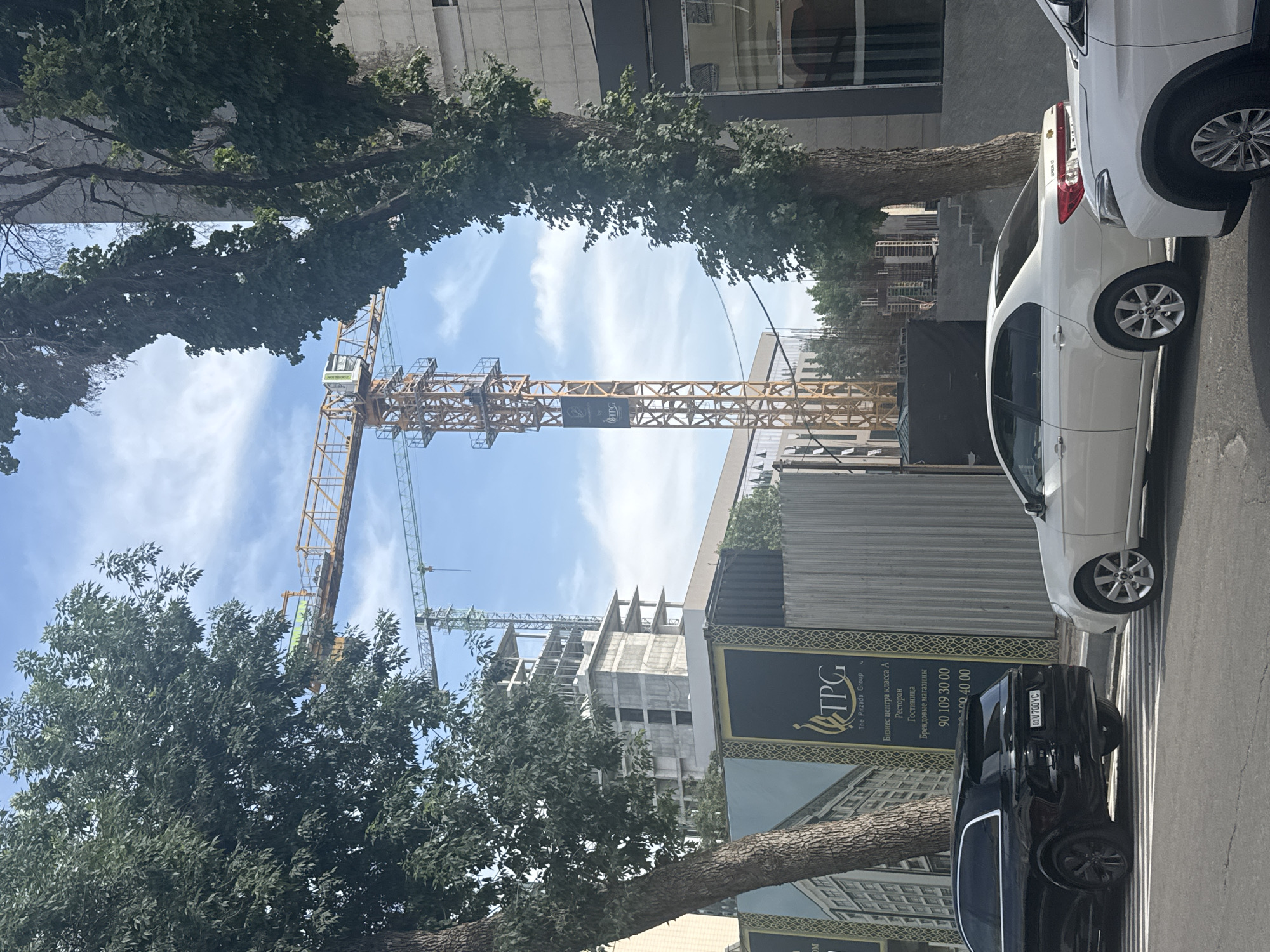 The image shows a street scene in Tashkent, Uzbekistan. On the right side of the frame, there are parked vehicles including a white sedan and a black SUV. A sidewalk runs along the right side. In the background, construction is taking place, with a building under construction visible alongside two construction cranes. A billboard with "TPG" (The Pirzada Group) is placed close to the cars, also displaying text in Russian. The sky is blue with some clouds. Trees are seen in the foreground, partially obstructing the view of the construction site.