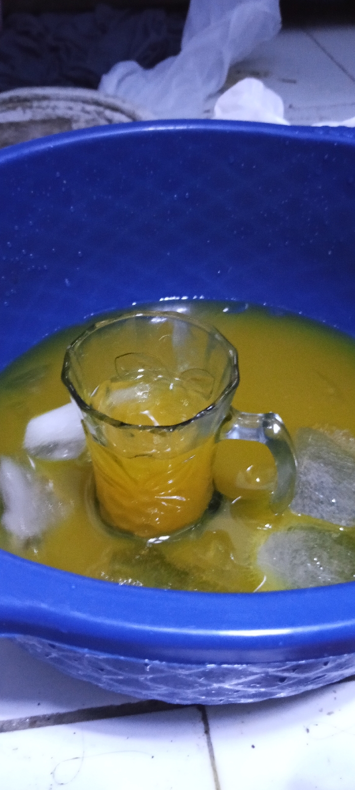 A blue plastic basin is filled with yellow liquid and ice. A glass pitcher containing yellow liquid sits in the center of the basin. The basin is sitting on a white tile floor. The location is Cangkuang, Indonesia, at coordinates -7.065 latitude, 107.552 longitude.