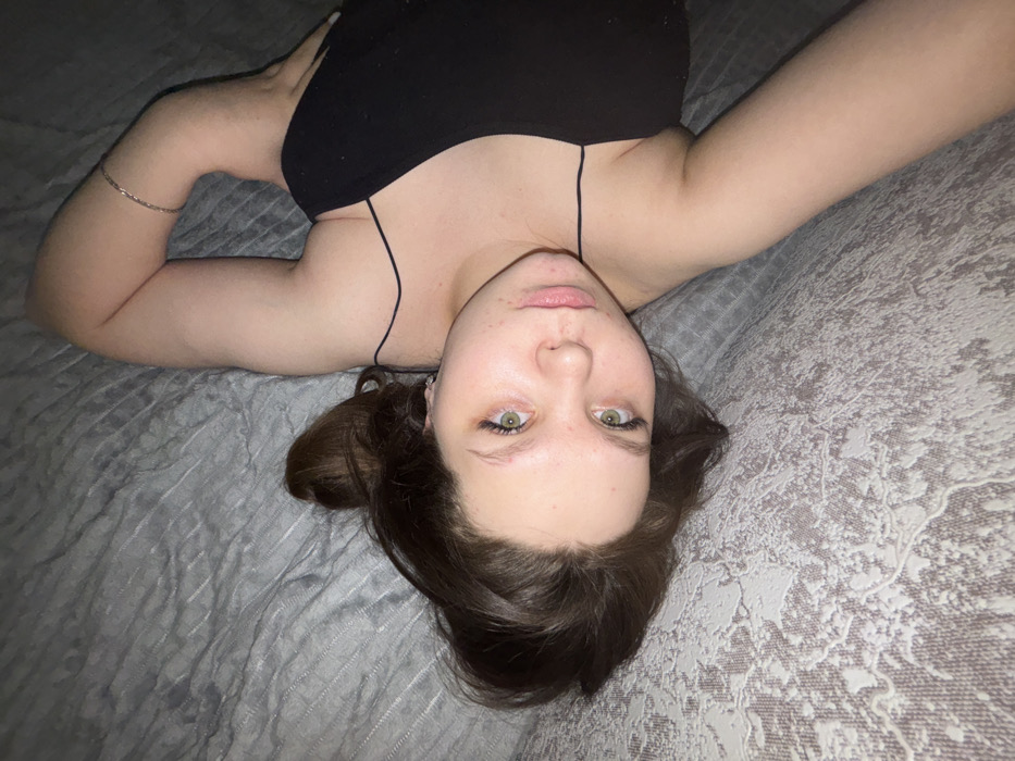 The image depicts a young adult female lying on her back, angled with her head towards the lower left of the frame, gazing directly at the camera. She has medium-length dark brown hair, green eyes, and light skin. She is wearing a black top with thin spaghetti straps. A silver-toned upper arm band is visible on her right bicep. Small earrings are discernible in her left ear. Her right arm is bent, with the hand out of frame; her left arm is mostly out of frame. The individual is positioned on a light grey or muted silver fabric surface featuring an intricate, raised, abstract or floral pattern, likely a bedspread or upholstered furniture. The lighting is soft and even, suggesting an indoor setting. The specific location within Perm, Russia, cannot be determined from the visual evidence presented in the image.
