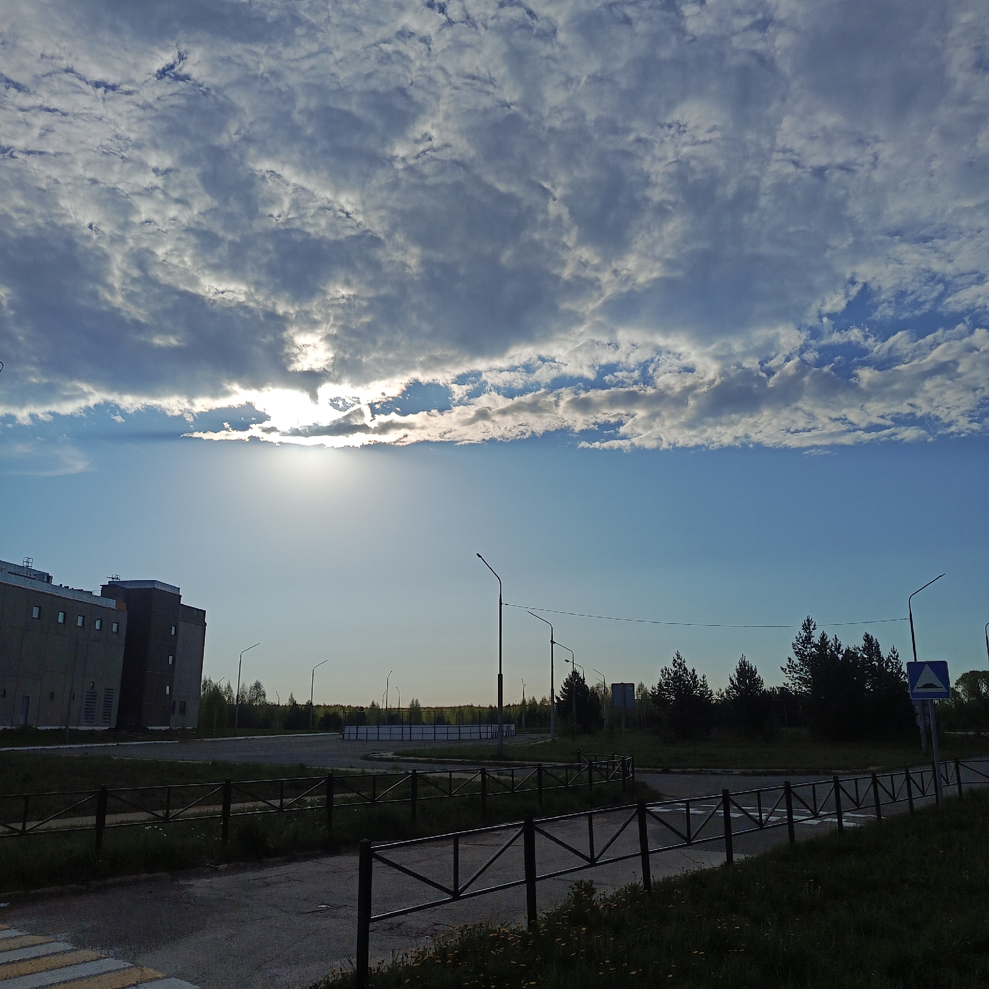 The image shows an urban scene in Марковский, Russia. The sky is mostly covered by clouds, with the sun shining through a break in the cloud cover. The sun is low on the horizon. In the background is a large, gray building with multiple windows. Streetlights are visible in the midground. A road runs across the bottom of the frame, marked with a pedestrian crosswalk. A black metal fence lines the side of the road. The foreground is dark, with grass and some yellow wildflowers. In the background a forest area can be seen.